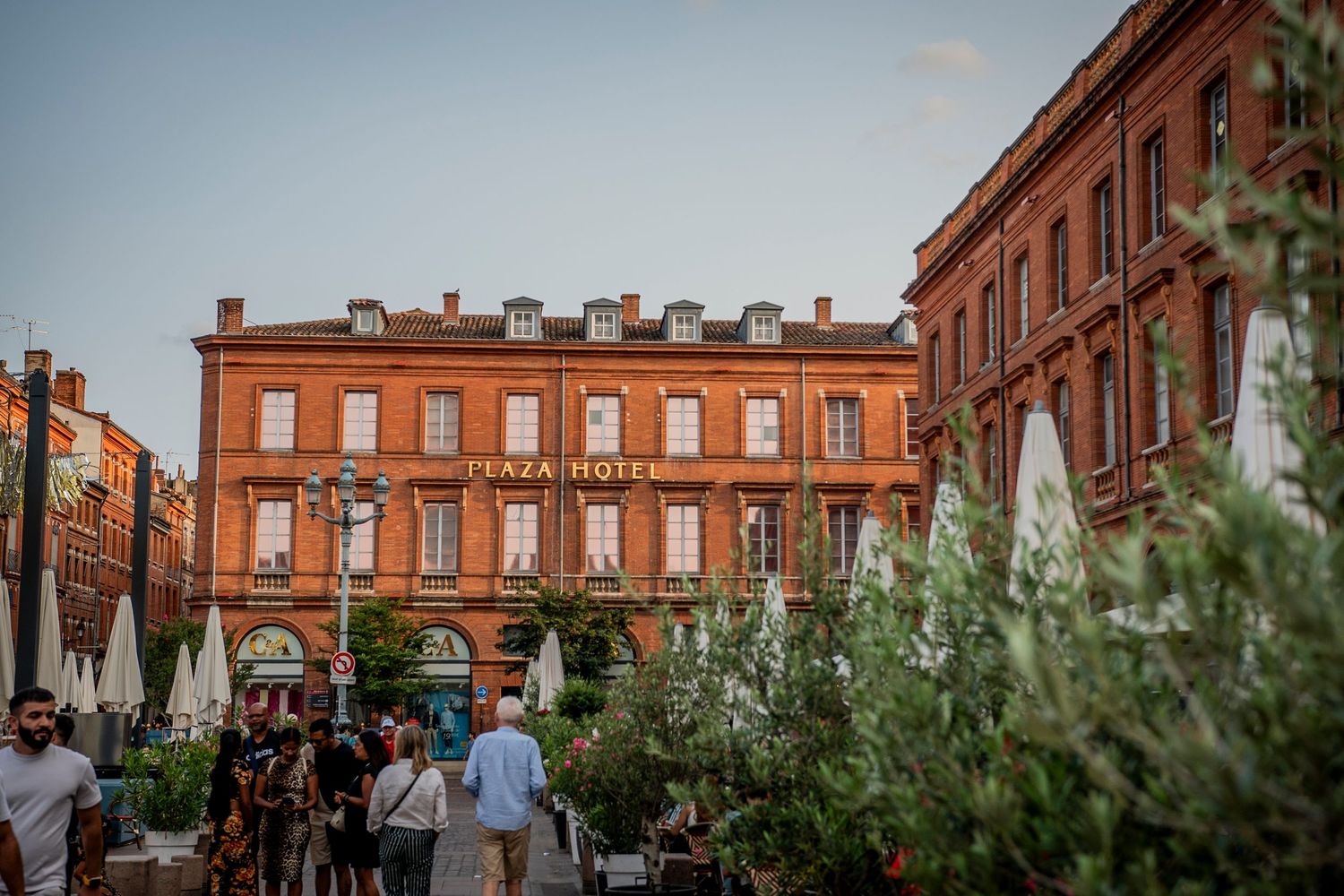 Red-brick colonial architecture building with pedestrians walking past shops and greenery in a historic square.