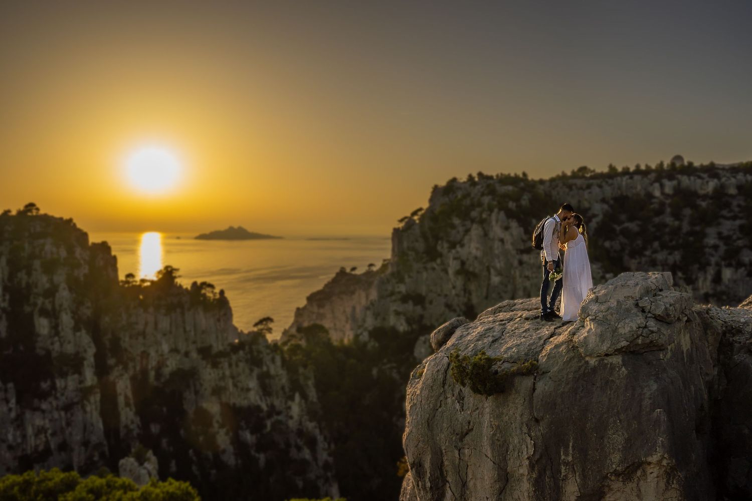 Couple in wedding attire standing on cliff edge during golden sunset.