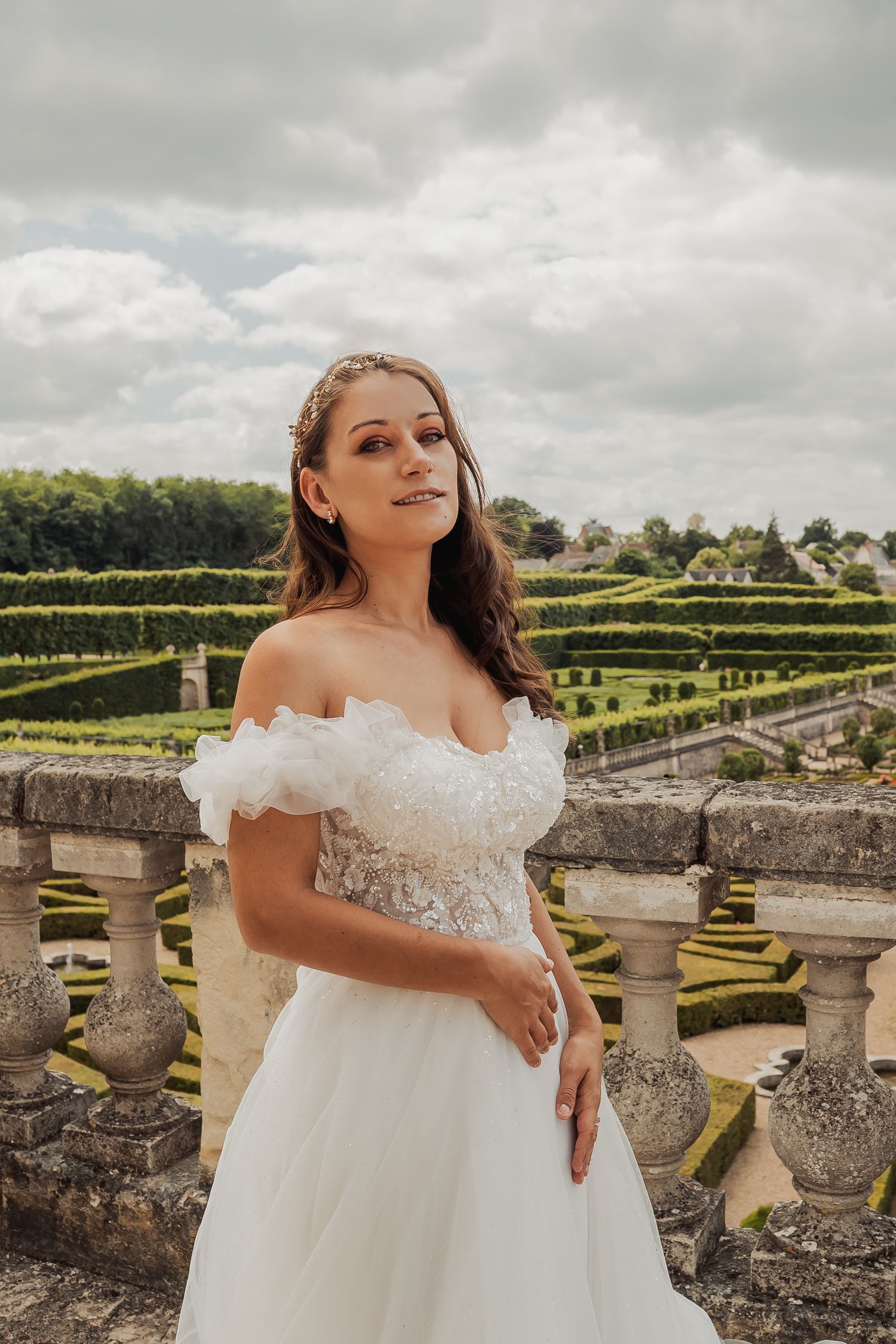 Portrait of bride in off-shoulder lace wedding dress standing on stone balcony overlooking formal garden landscape.