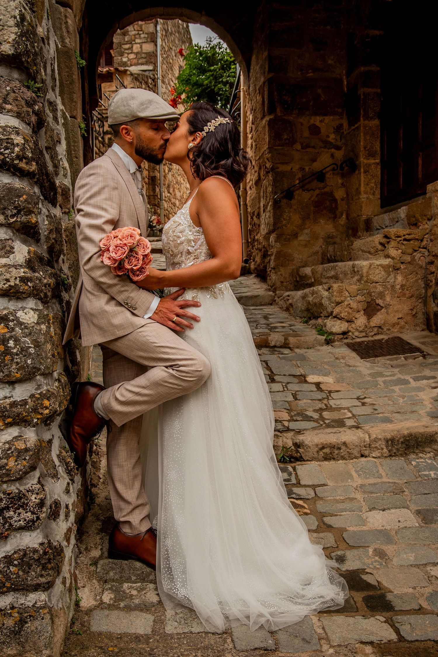 Wedding couple embraces on rustic cobblestone street with stone walls.
