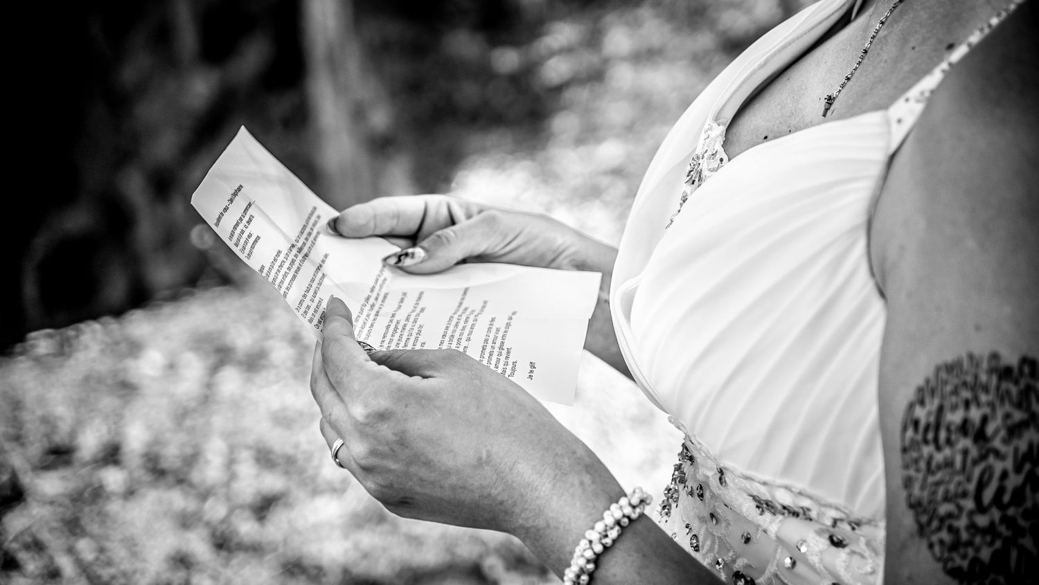 Person in white dress reads wedding vows from paper during outdoor ceremony.
