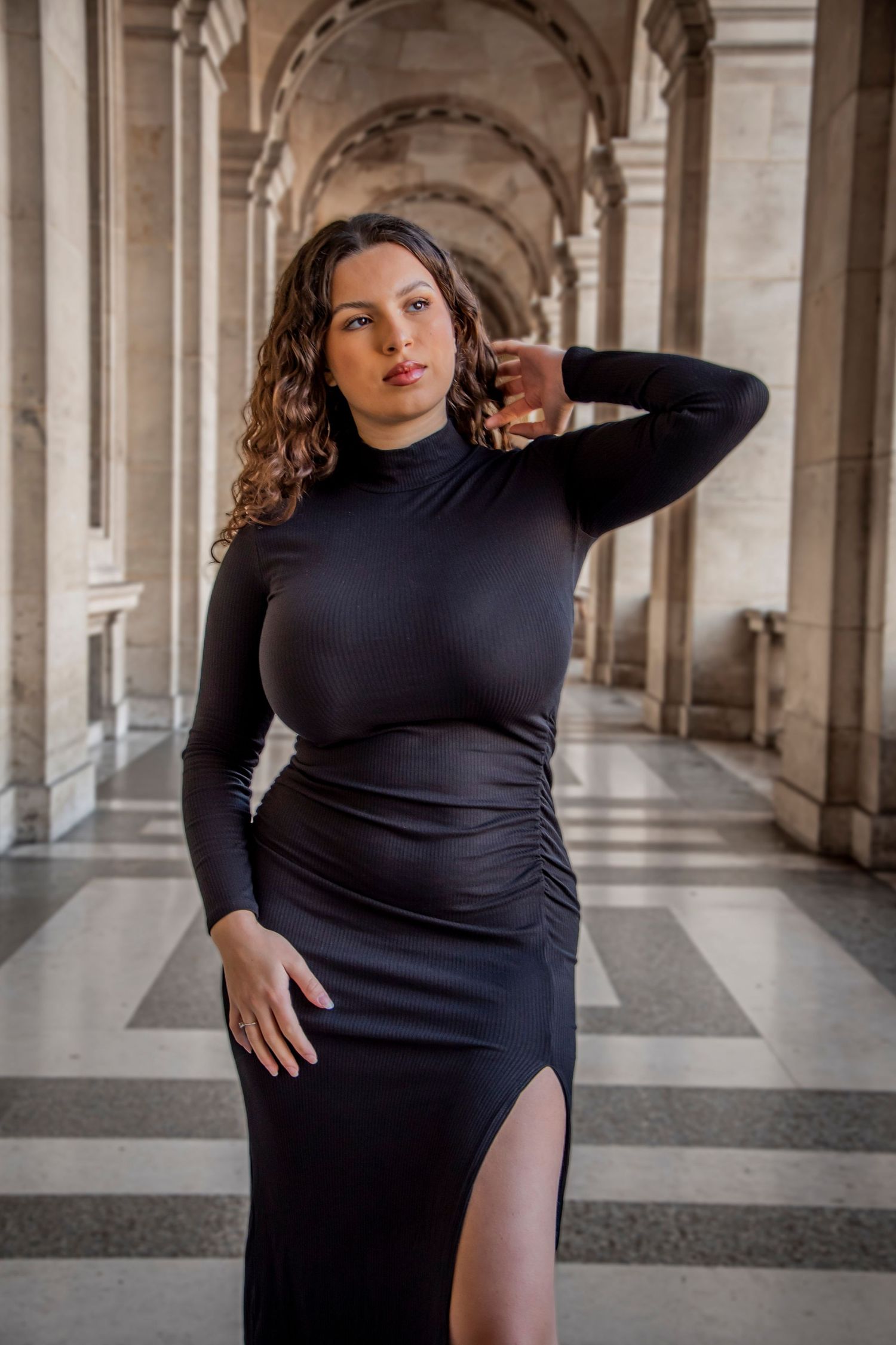 Person in black dress poses elegantly in arched colonnade with dramatic lighting and architectural details.