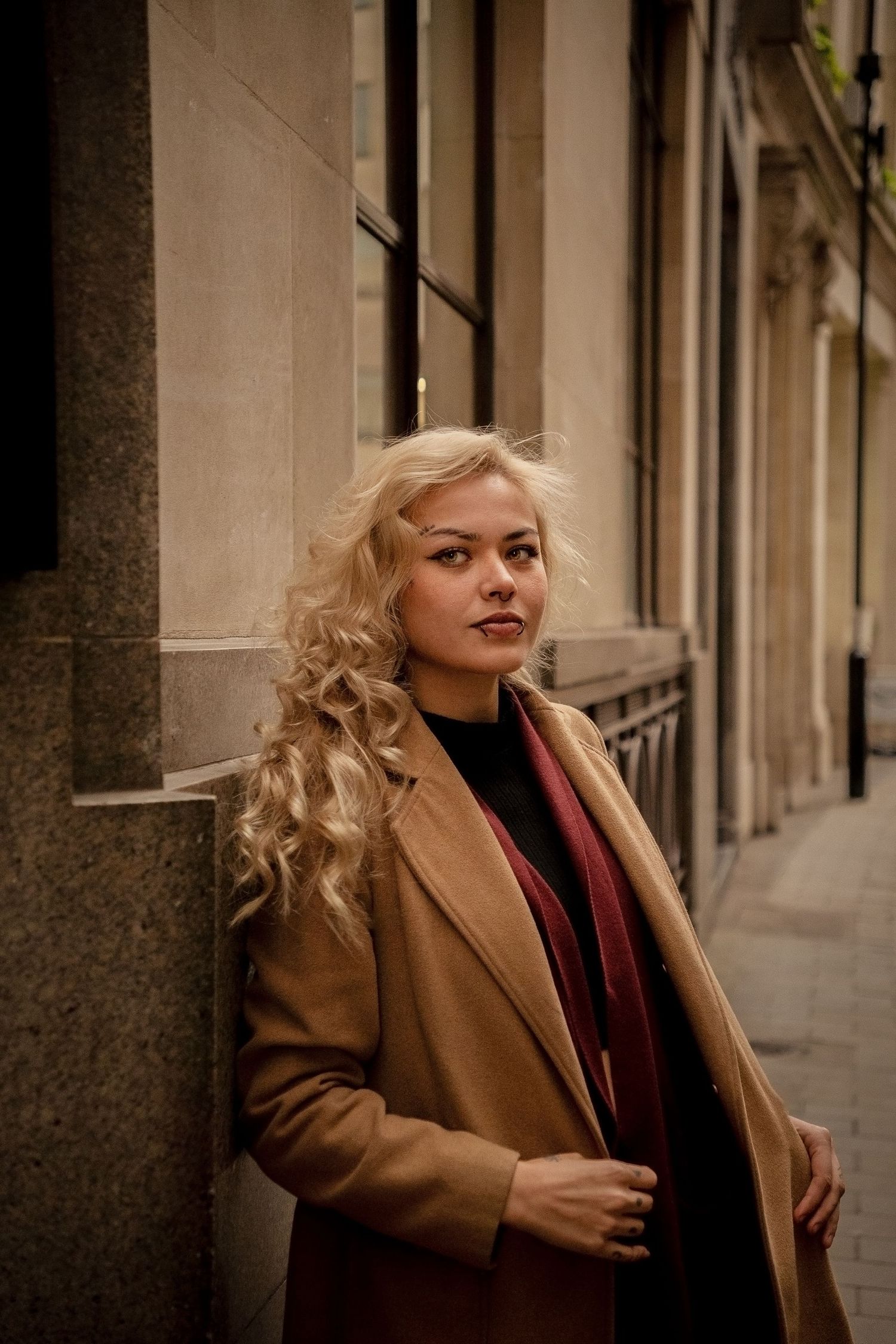 Woman in camel coat with blonde curly hair poses against urban building backdrop.