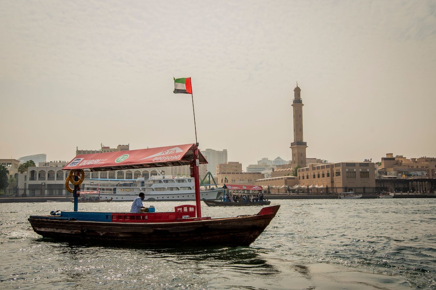 Traditional wooden abra boat cruises Dubai Creek with minaret of Grand Mosque visible in background under hazy sky.