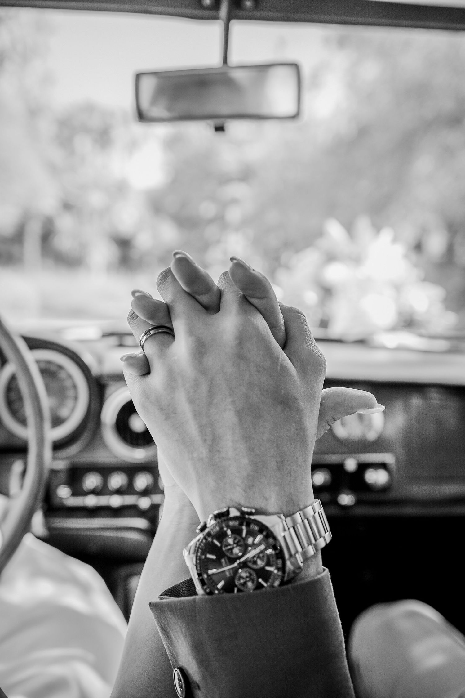Black and white photo of clasped hands with luxury watch on car steering wheel.