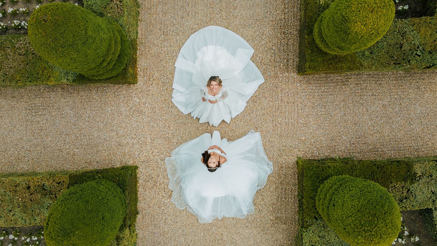 Aerial drone view of a couple in white wedding dresses creating a symmetrical pattern on gravel path surrounded by manicured lawn.