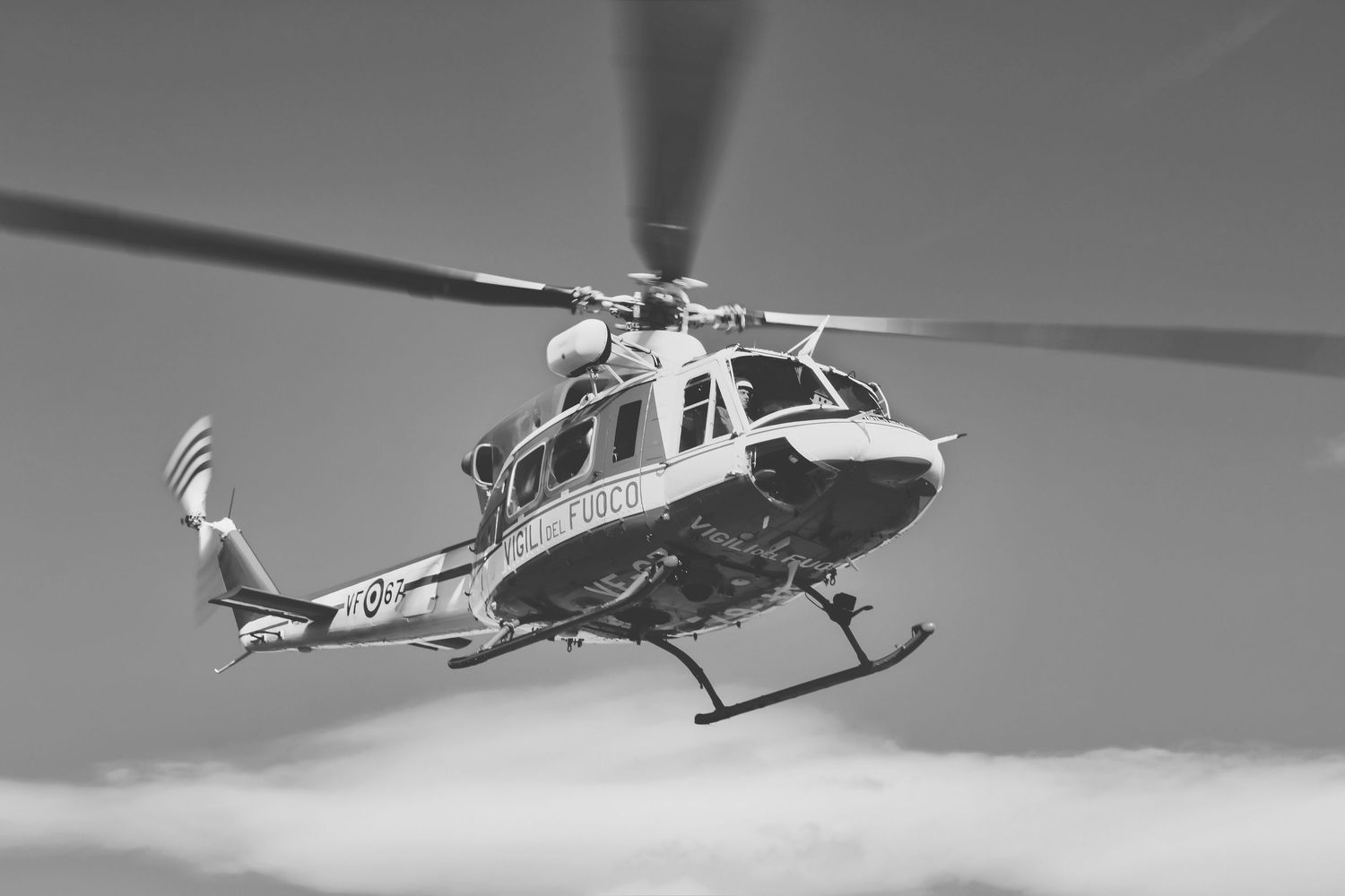A Bell 412 helicopter hovers in mid-flight against a cloudy sky in black and white photography.