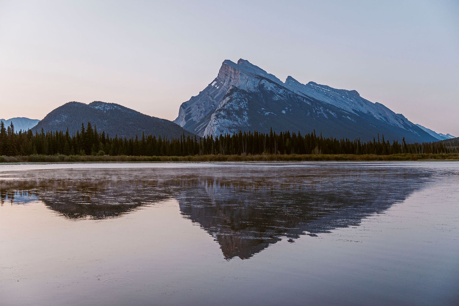 Panoramic mountain landscape reflected in calm lake waters under dramatic cloudy sky at sunset in Canadian wilderness.