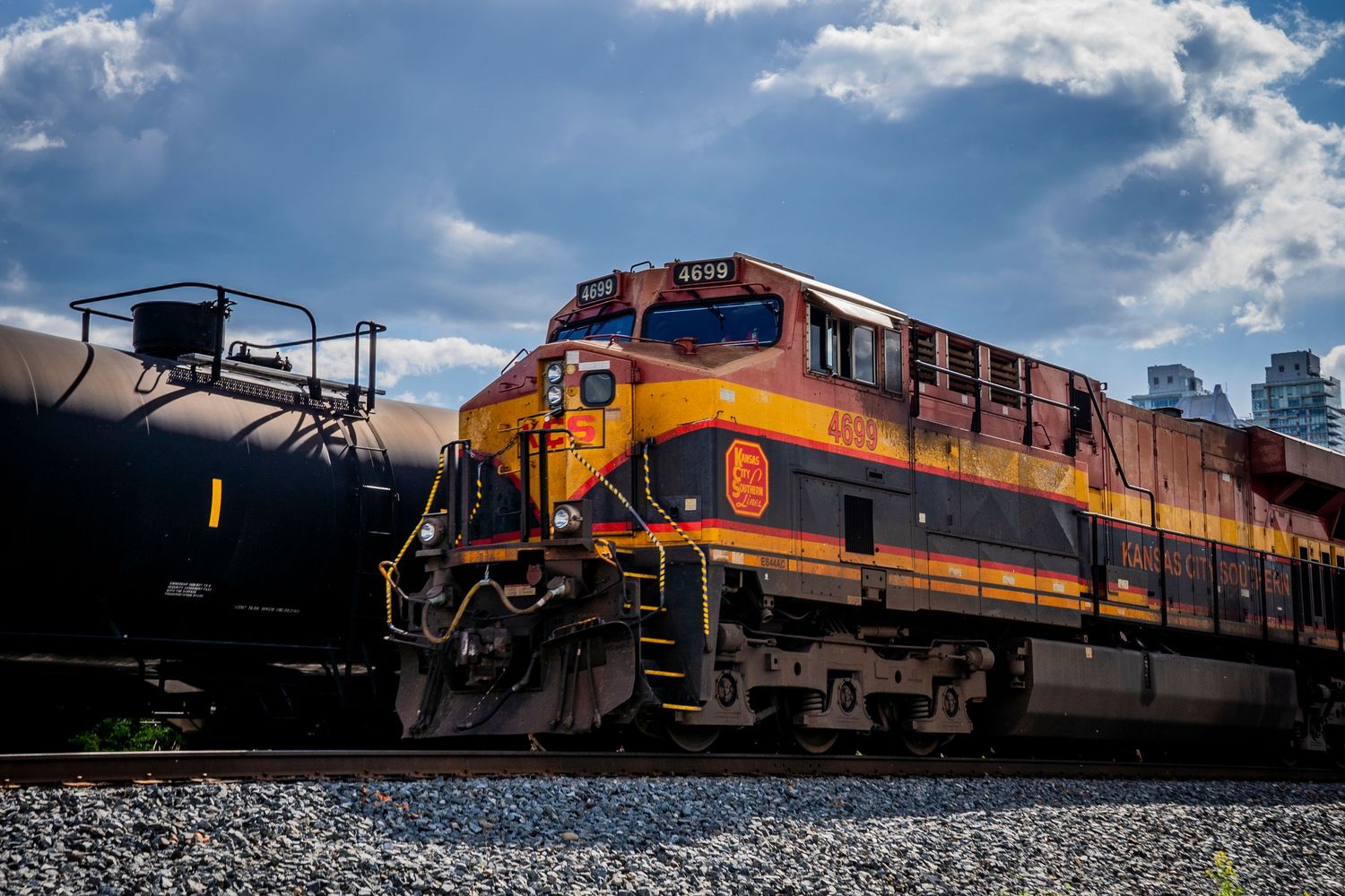 A Kansas City Southern locomotive in orange and black paint scheme sits on railroad tracks under dramatic cloudy skies.