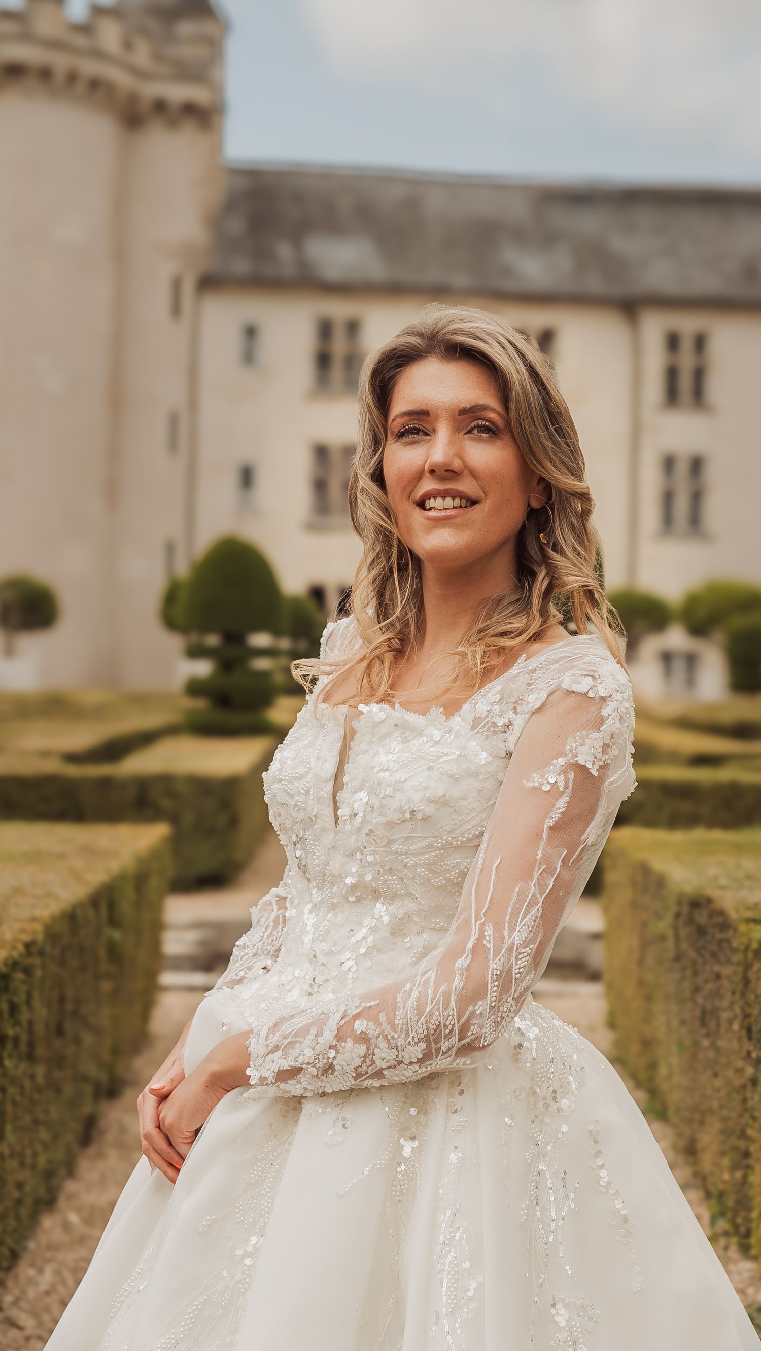 A lovely bride in a detailed white lace wedding gown poses gracefully in front of a manicured garden hedge.