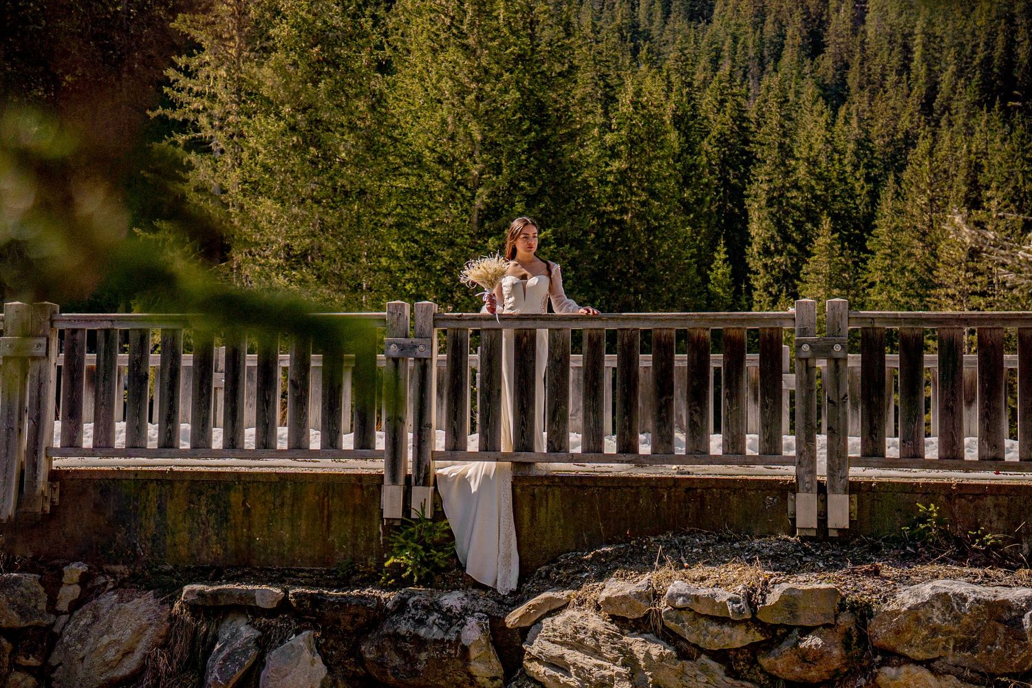 A couple stands on a rustic wooden bridge overlooking a mountain stream surrounded by evergreen forest.