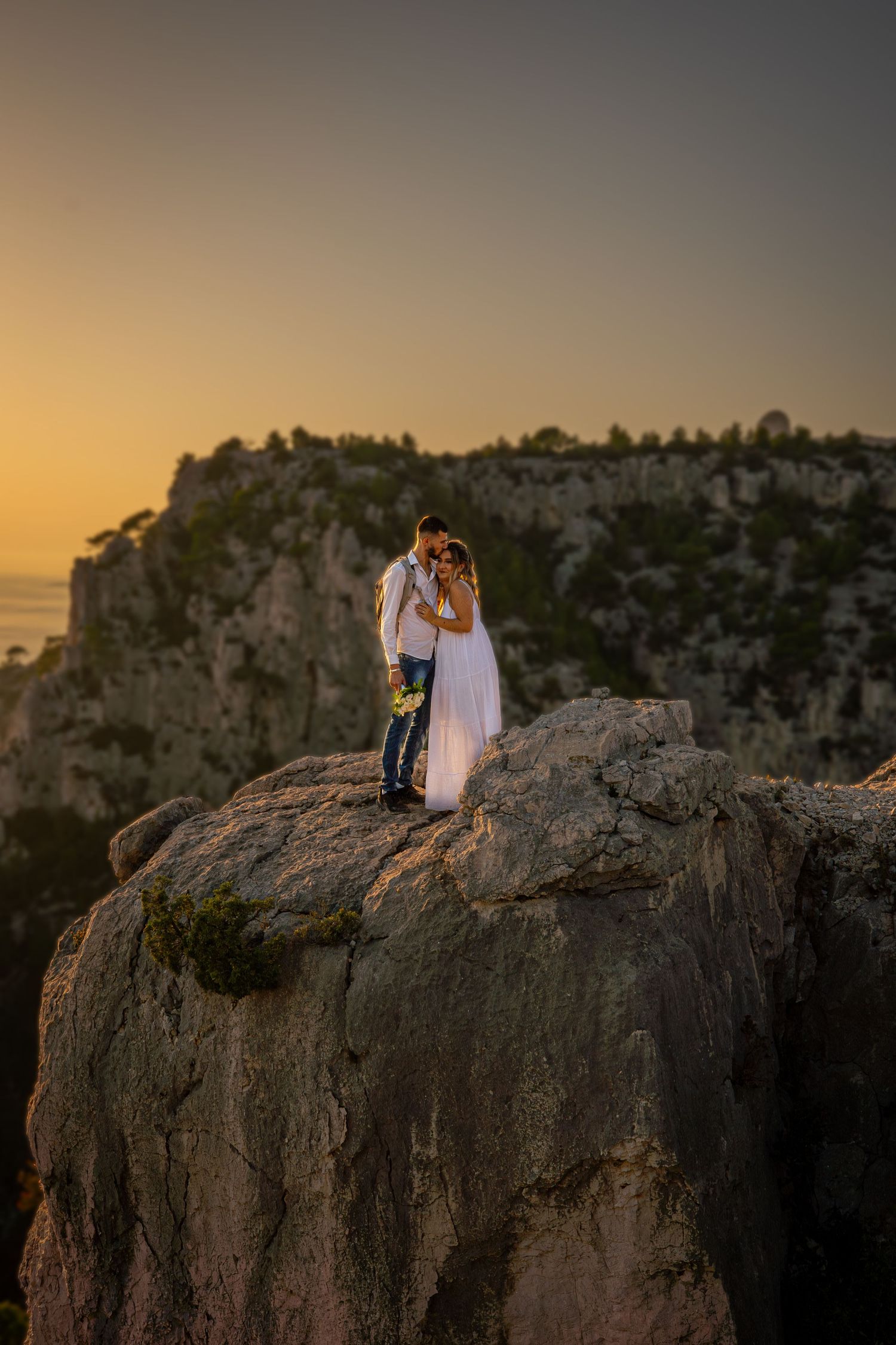 Two figures embrace on a dramatic cliffside overlook during golden hour sunset with mountains in background.