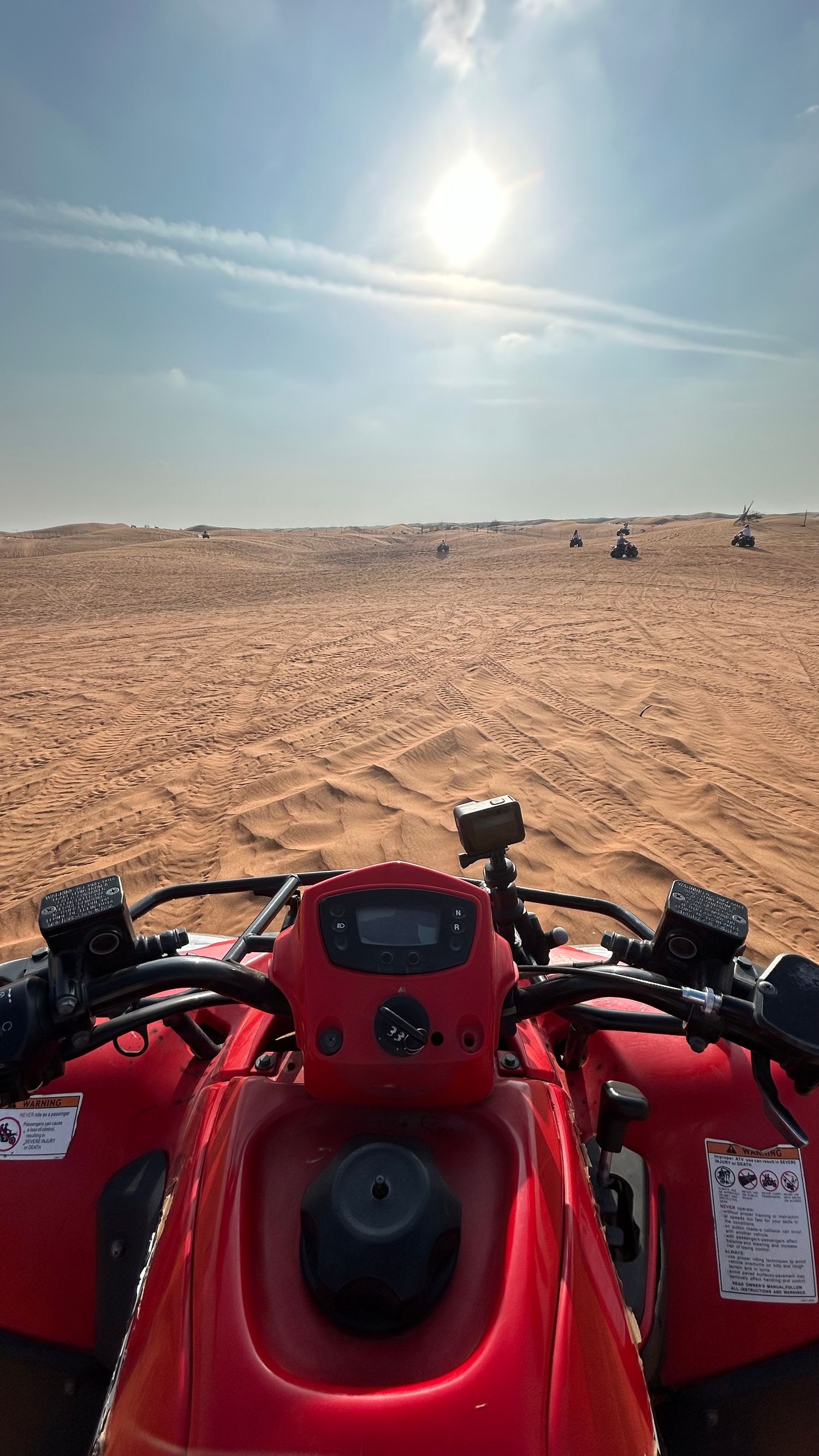 Red ATV handlebars and controls shown against a sunny desert landscape with rolling sand dunes stretching to the horizon.