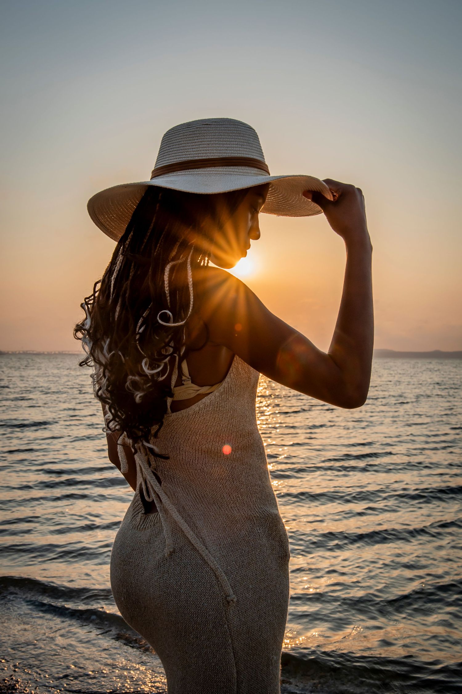 A silhouetted figure in a hat stands against a sunset over the ocean, creating a backlit atmosphere.