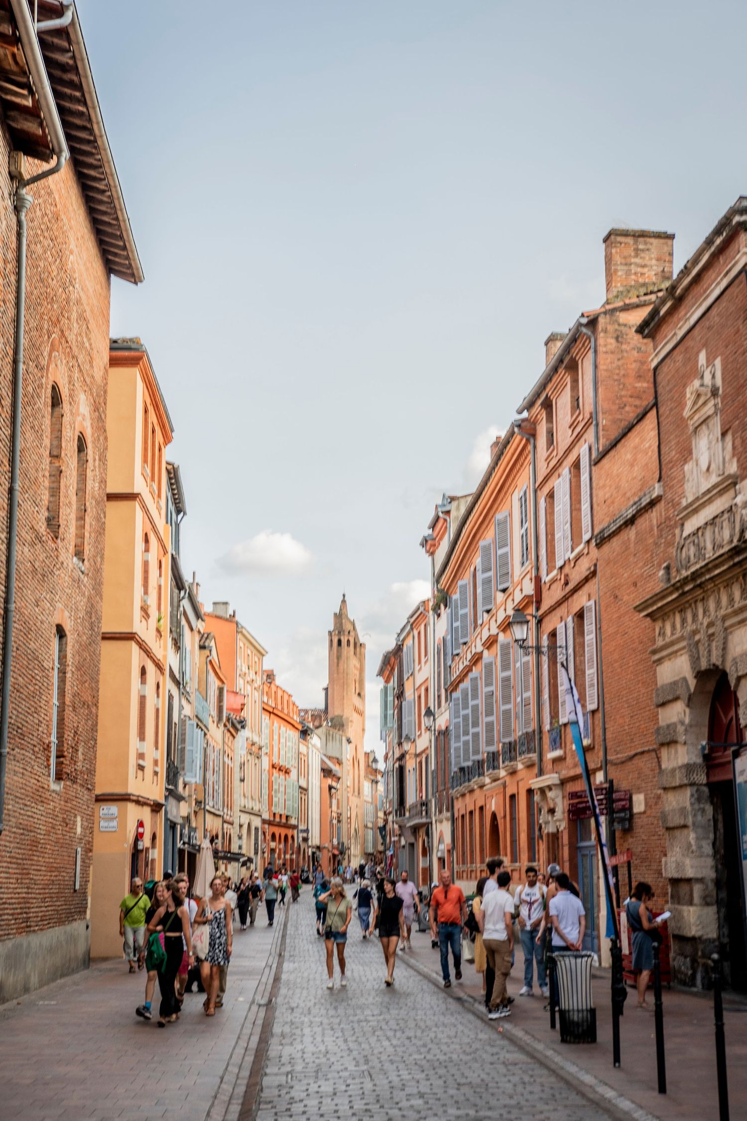 Historic cobblestone street in Toulouse, France with red brick buildings and people walking along a narrow pedestrian way.