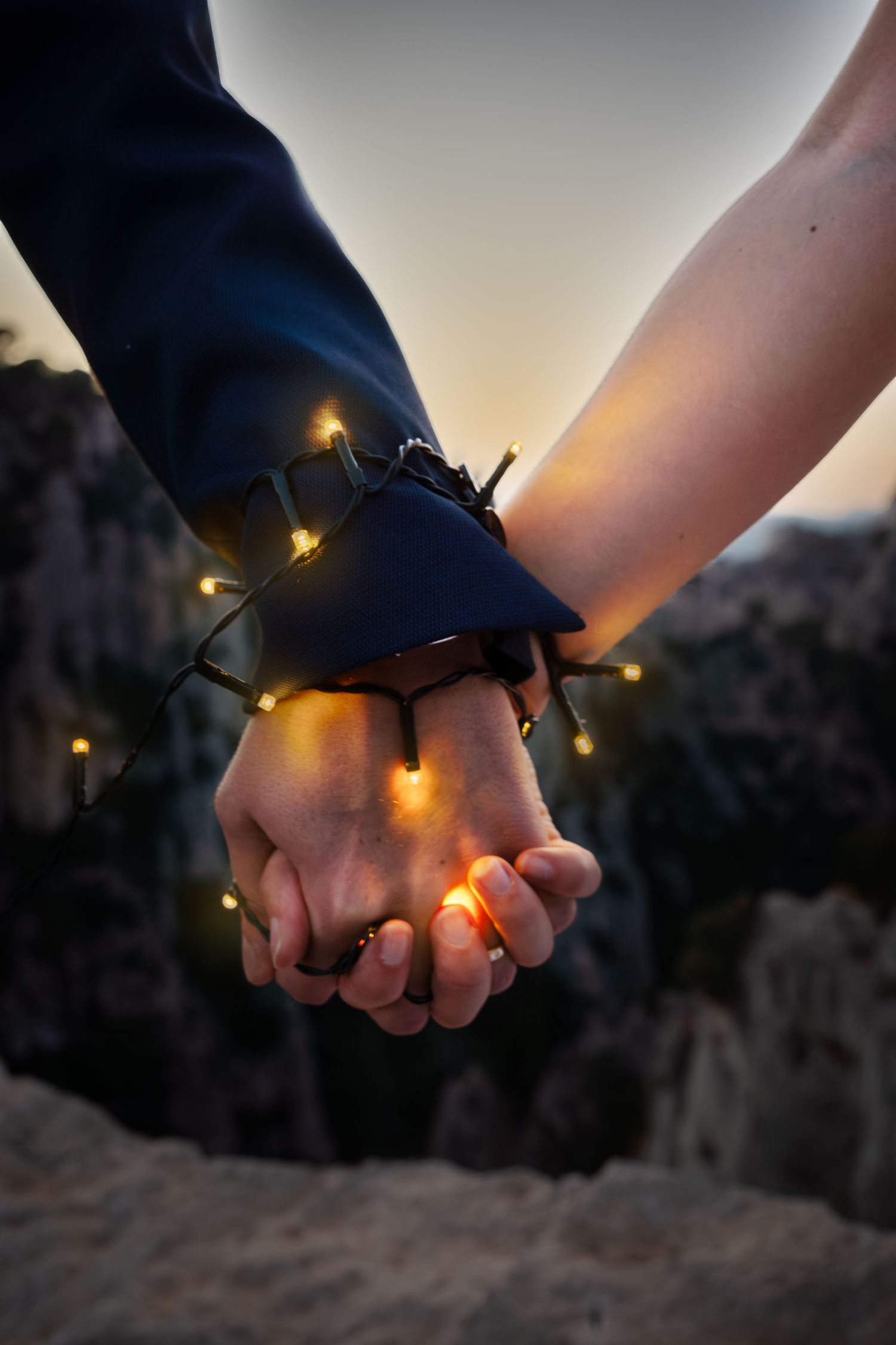 Two hands gently clasped together at sunset with twinkling string lights wrapped around them creating a romantic atmosphere.