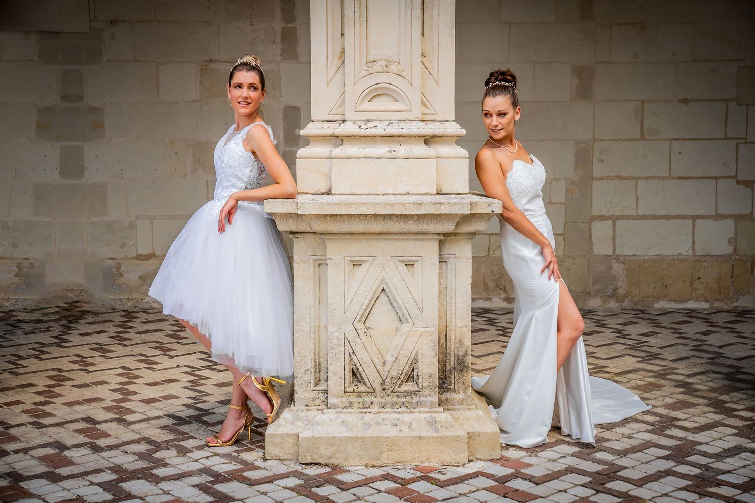Two dancers in elegant white dresses pose gracefully against an ornate stone pillar on a patterned floor.