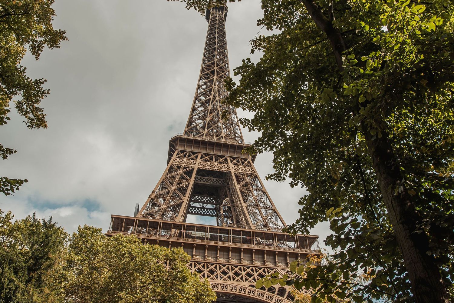 View of the iconic Eiffel Tower in Paris framed by green trees against a cloudy sky.
