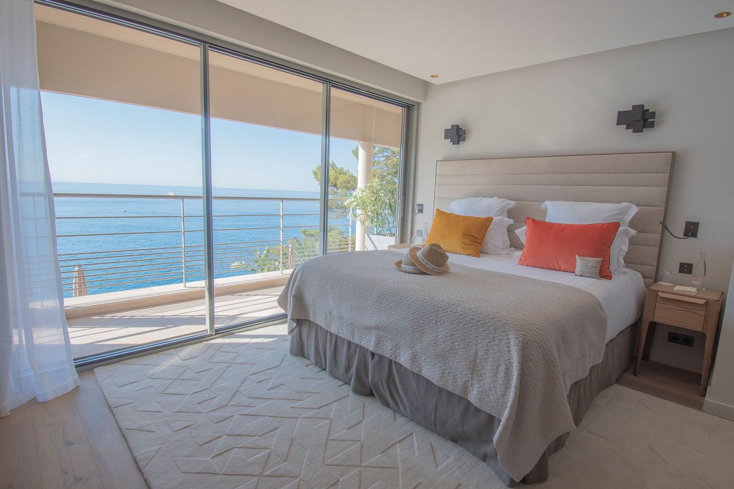 Modern coastal bedroom featuring crisp white bedding, coral pillows, and large sliding doors with panoramic sea views.