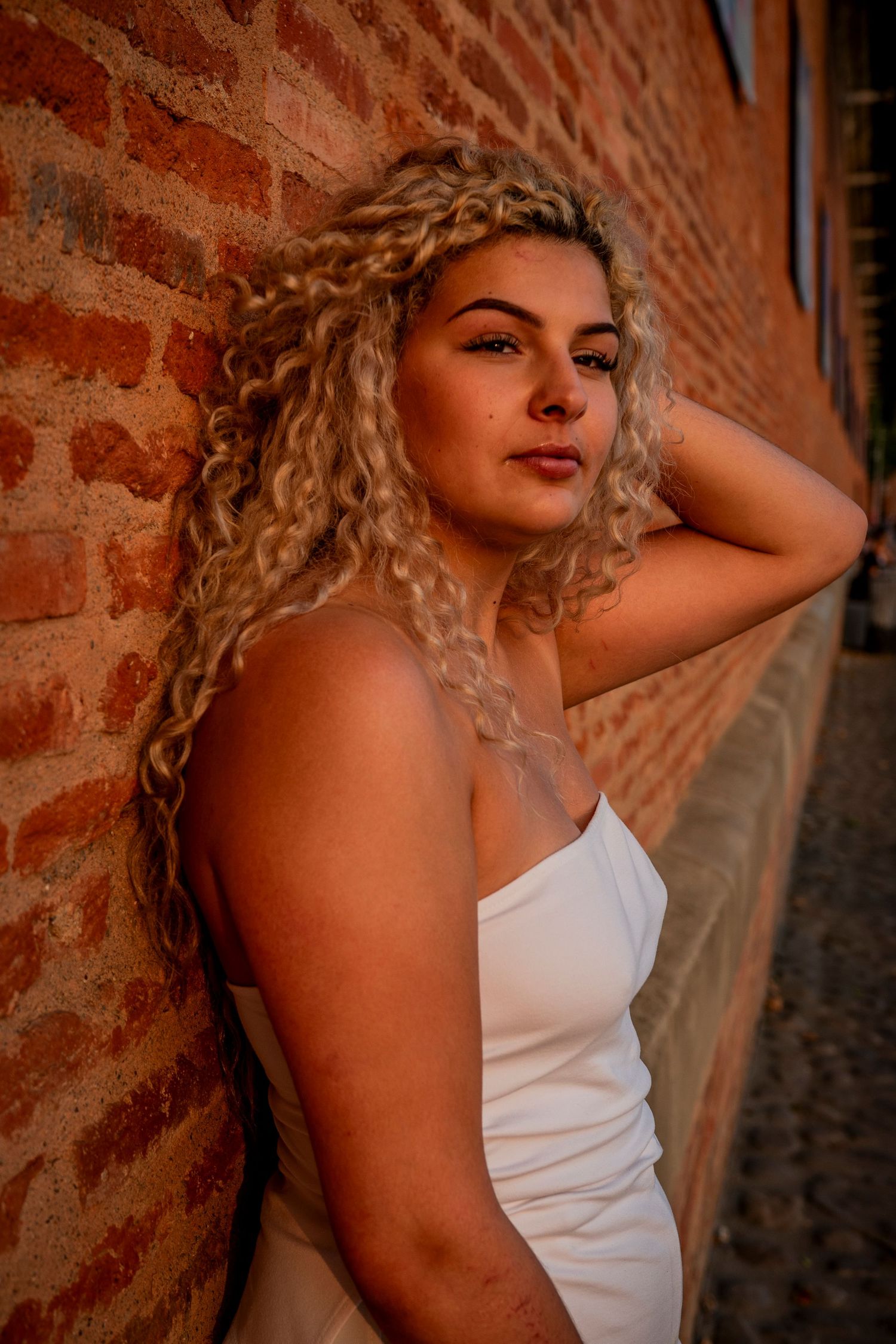 Person with long curly blonde hair poses in white dress against rustic brick wall in warm sunset lighting.