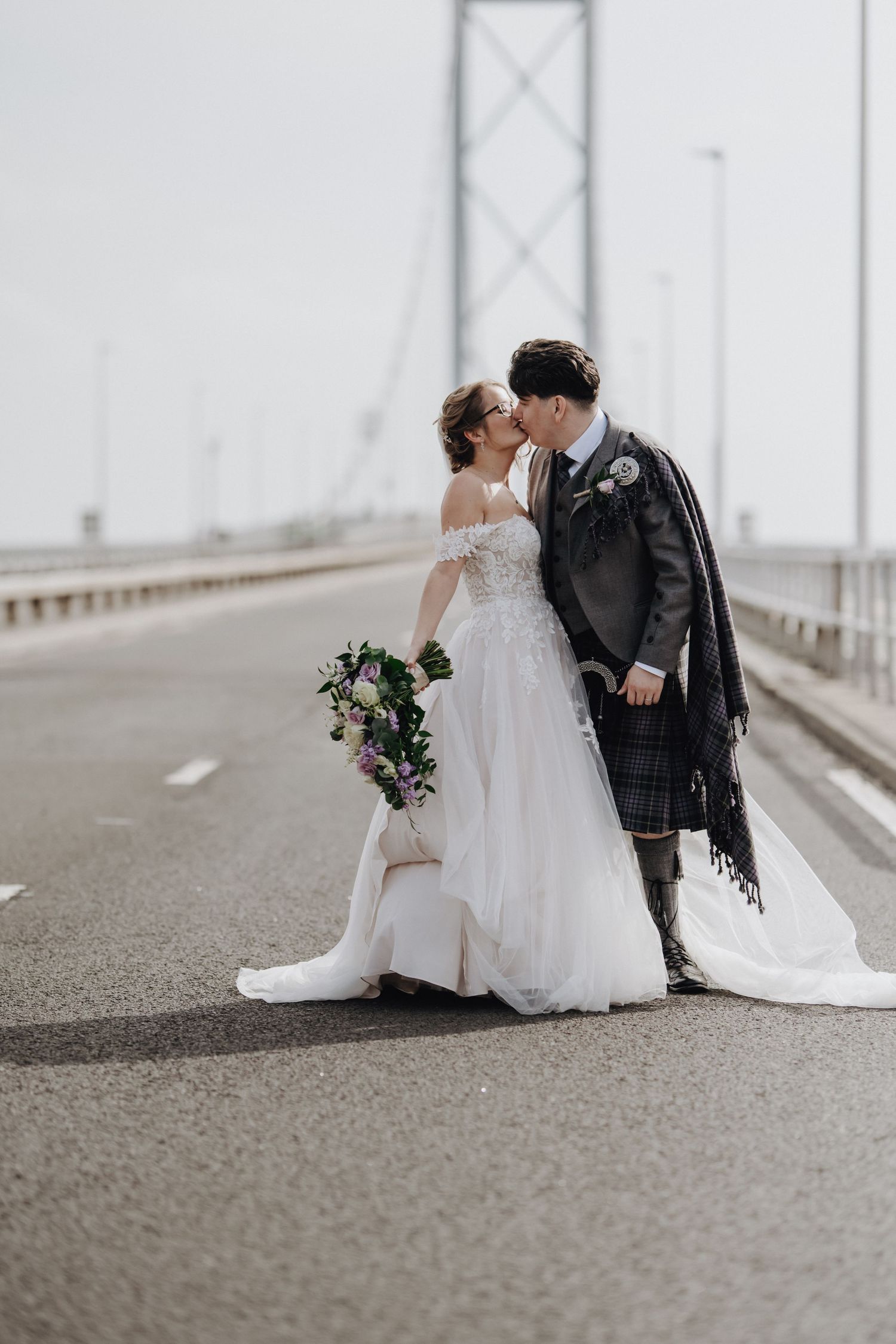 Romantic wedding couple sharing a moment on a misty bridge with the bride in white and groom in traditional kilt.