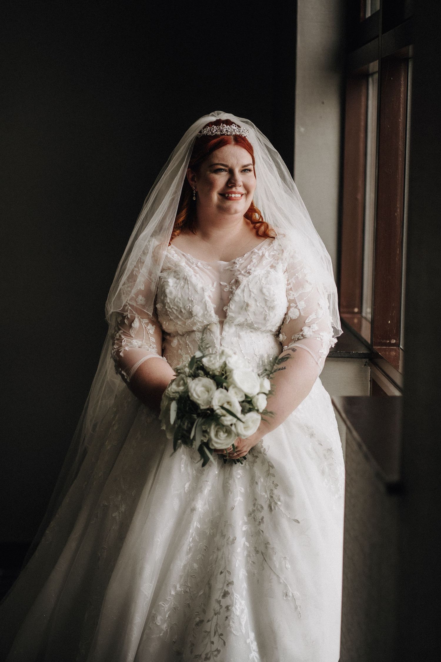 A bride in a beautiful lace wedding dress with sleeves poses by a window holding a white floral bouquet.