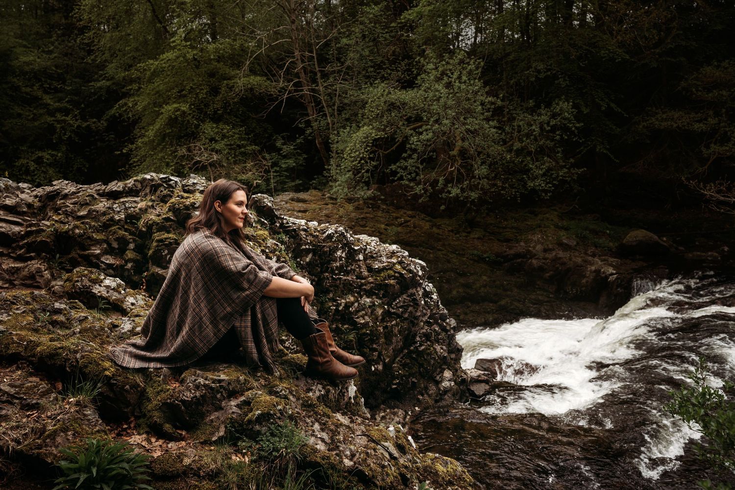 A person in casual clothing sits contemplatively on rocks beside a rushing waterfall in a lush forest setting.