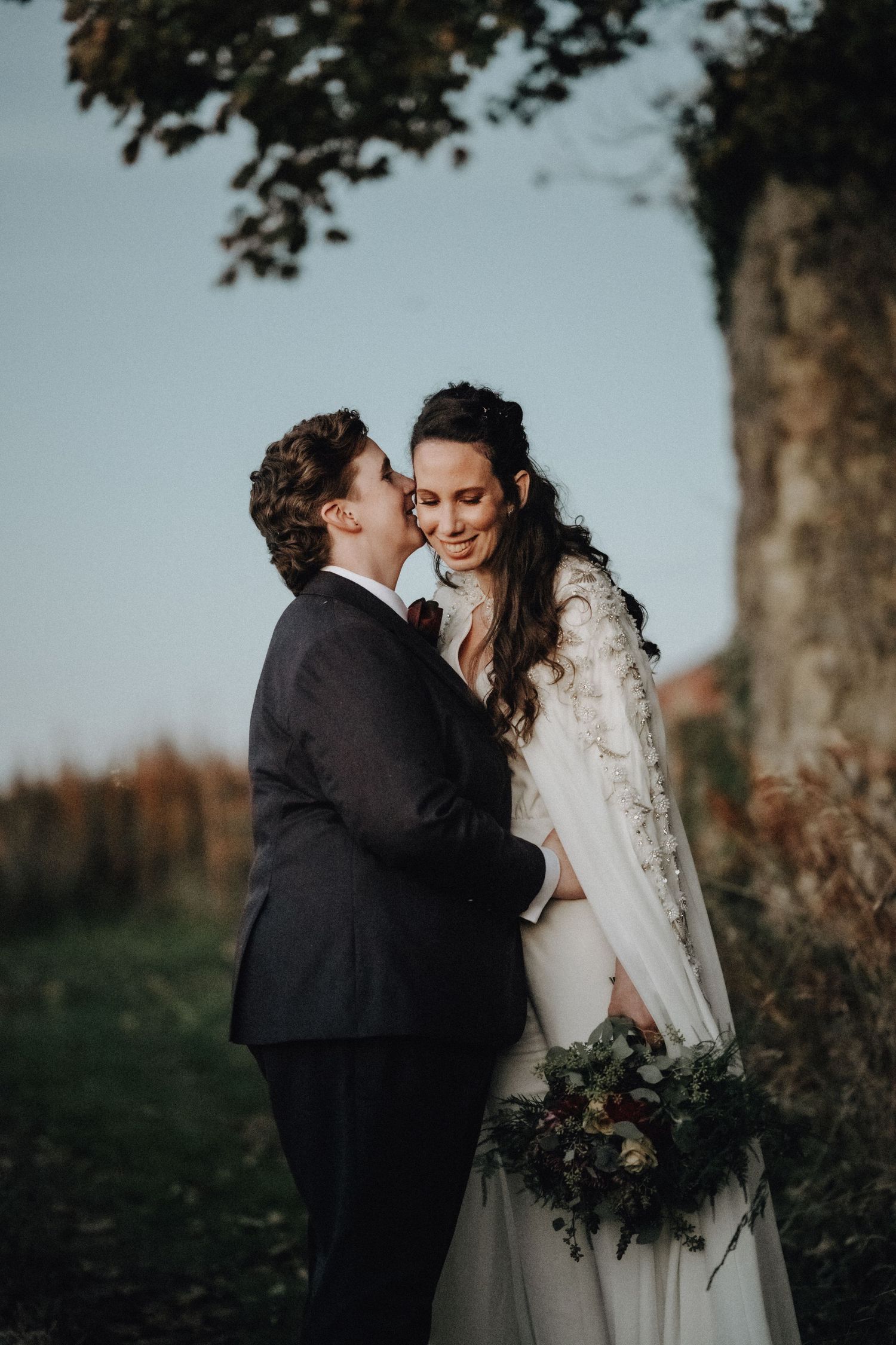 A romantic wedding portrait of a couple sharing an intimate moment under a tree at dusk.