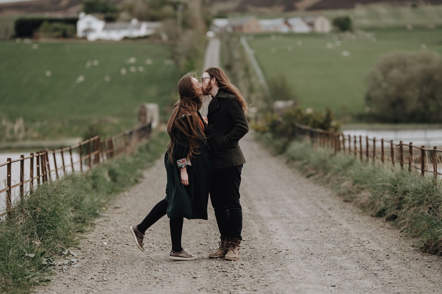 A couple shares a romantic moment on a countryside path with rolling hills and farmland in the background.
