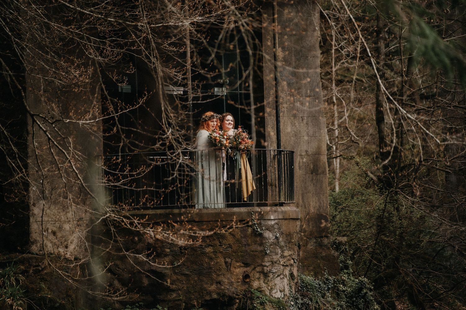 A romantic woodland wedding couple poses on a rustic wooden balcony surrounded by forest trees at dusk.