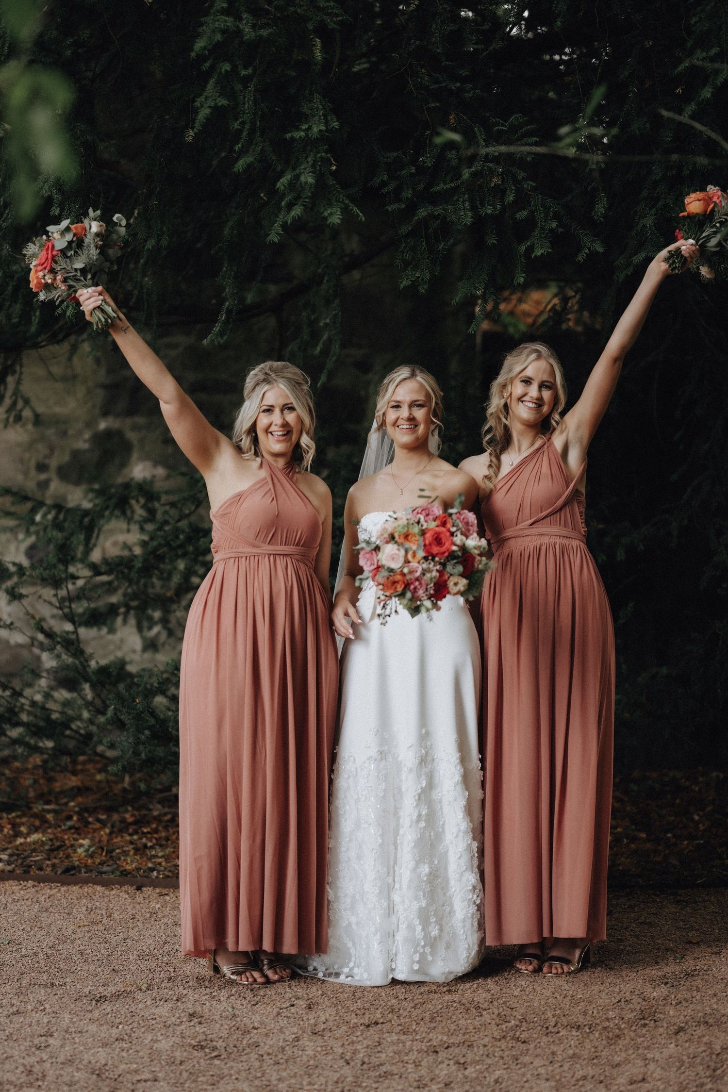 Three women in dusty rose dresses celebrate joyfully while holding floral bouquets against a stone wall.
