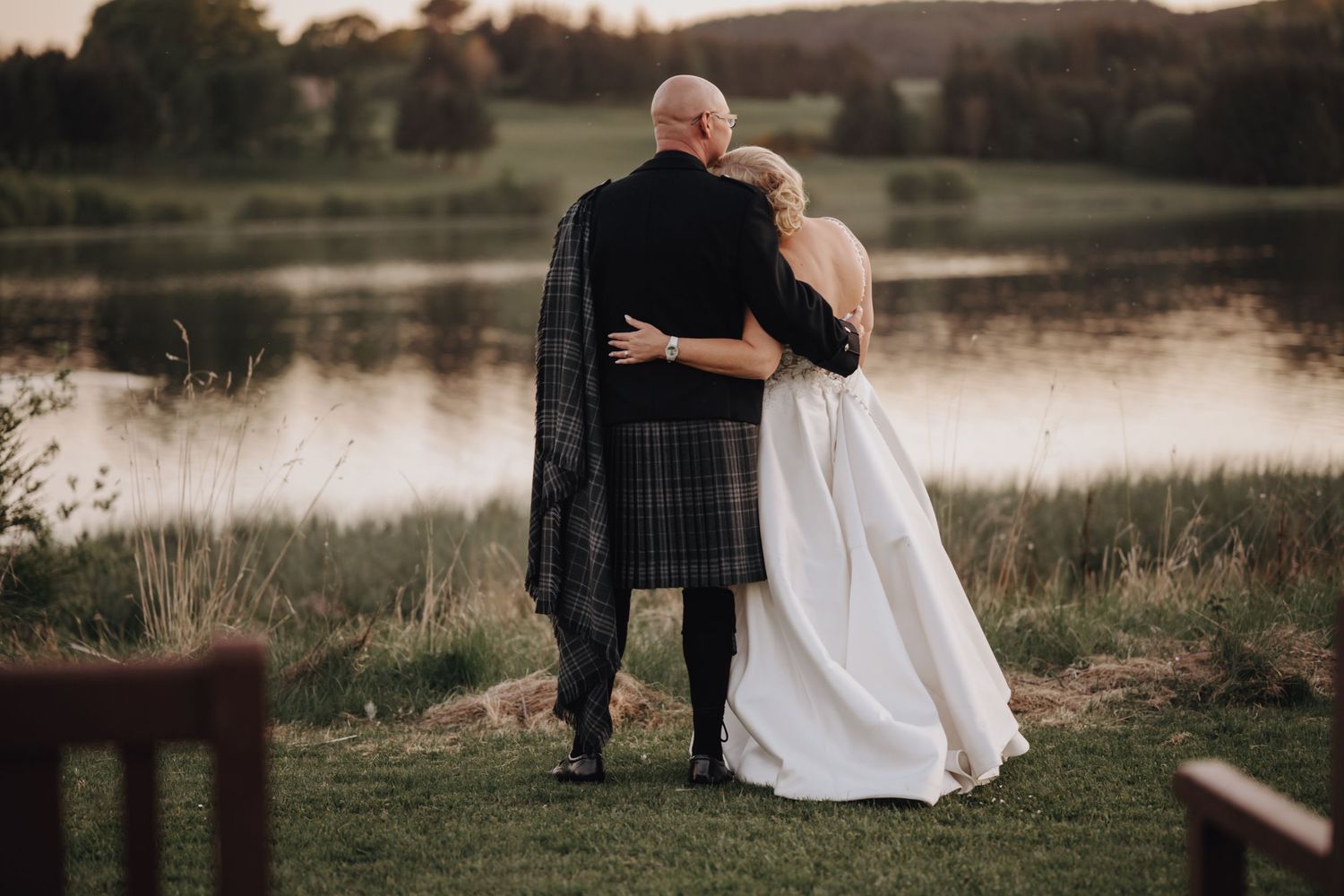 A couple in wedding attire embraces by a serene lake at sunset, with the groom wearing a traditional kilt.