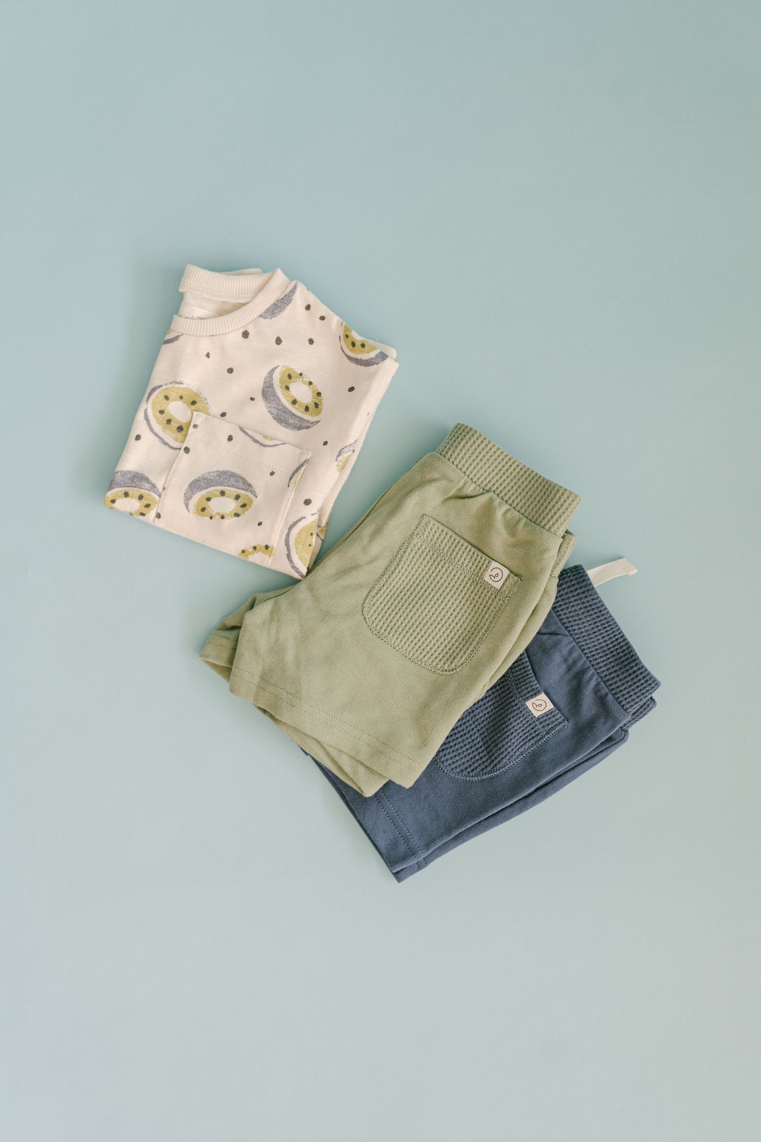 Folded baby clothes featuring a moon print onesie and sage green pants on blue background.