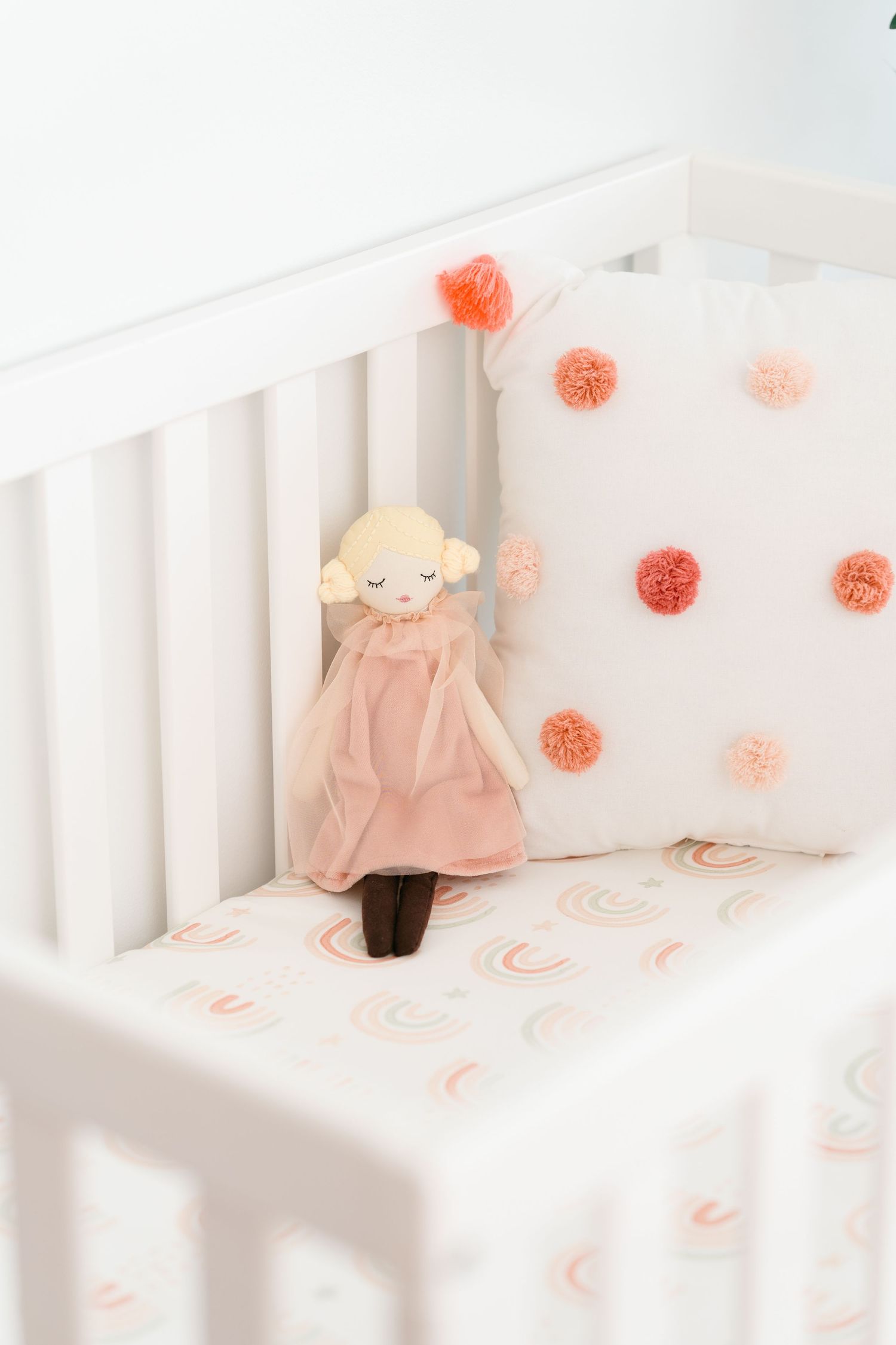 White crib with soft plush doll and coral pom pom decorations.