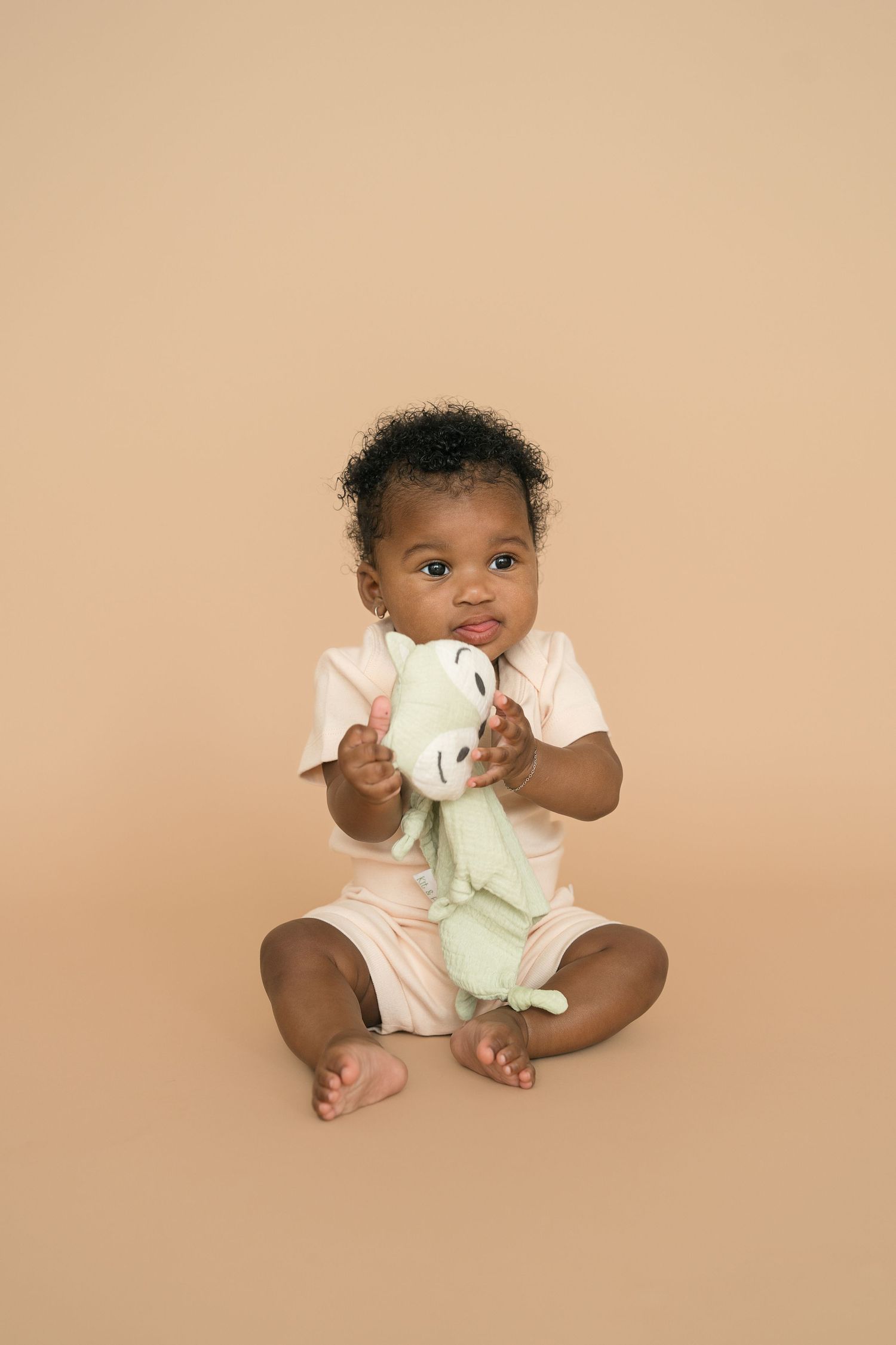 A small child wearing a light-colored outfit sits on a beige background holding a cloth.