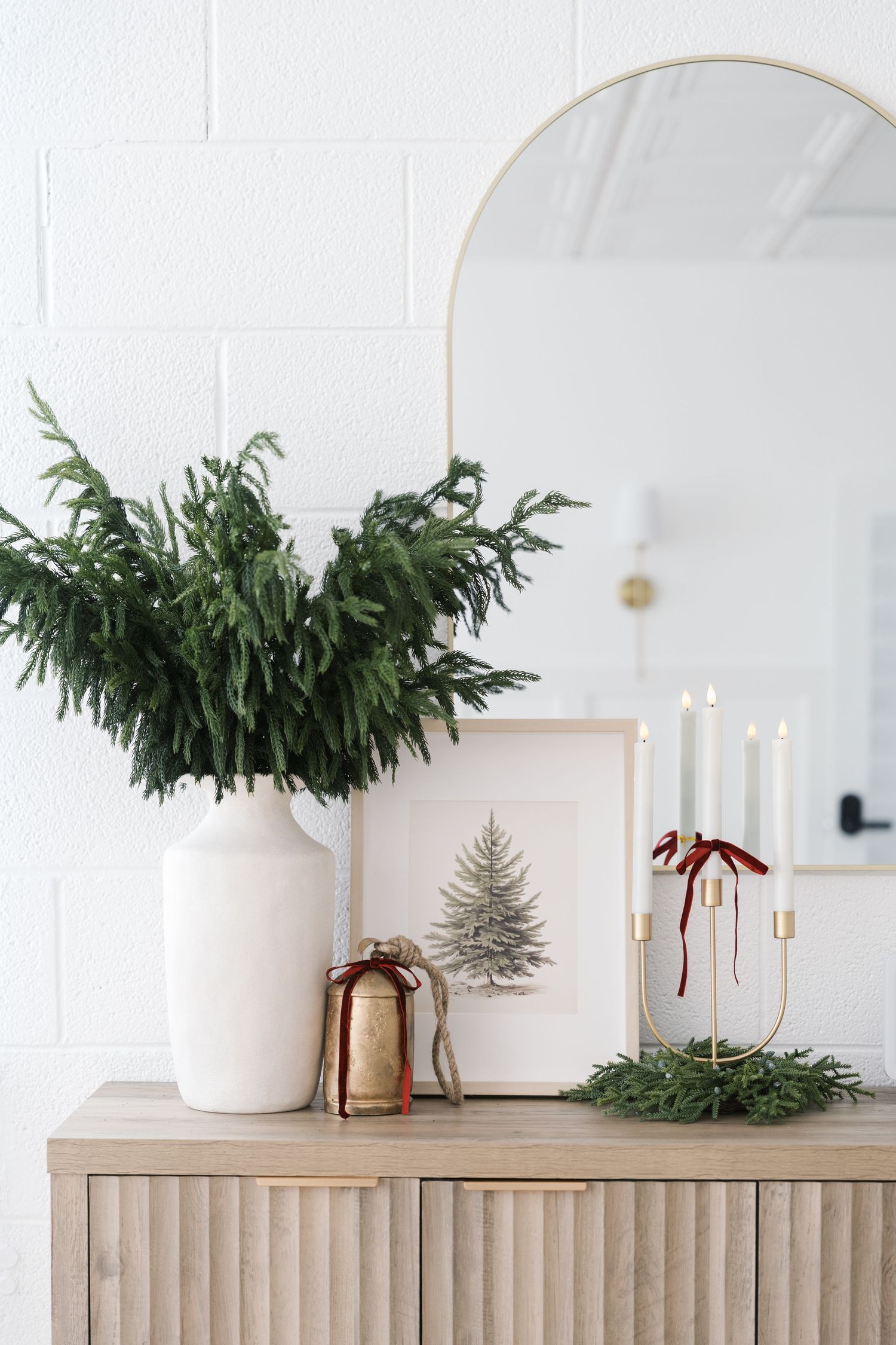 Minimalist holiday decor featuring evergreen branches in a white vase, candles and tree artwork on a wooden cabinet.