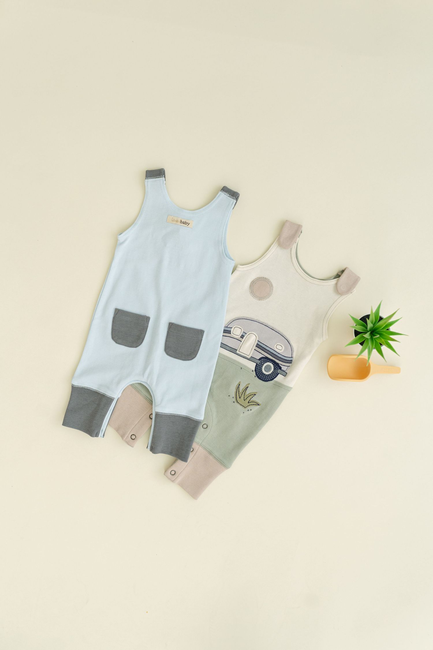 Two baby rompers with cute designs and a small potted plant displayed on beige background.
