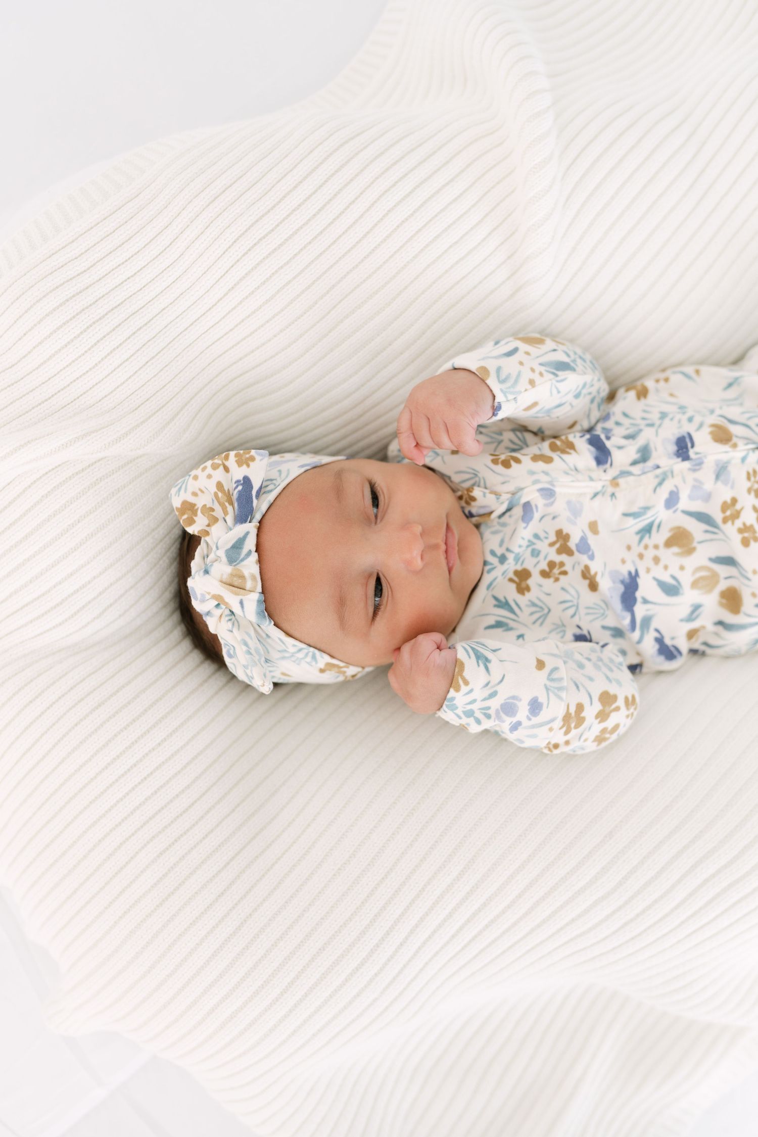 A baby wearing a floral outfit lies on white textured bedding.