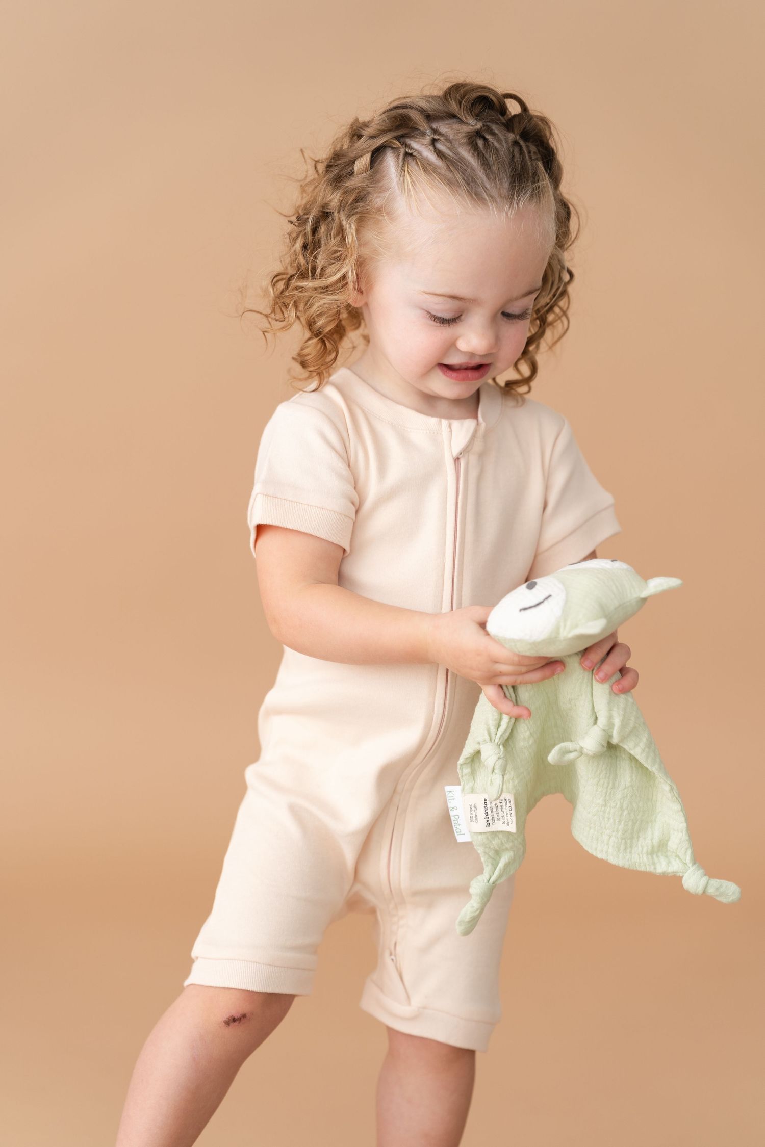 Child in cream colored romper playing with mint green plush toy against beige backdrop.
