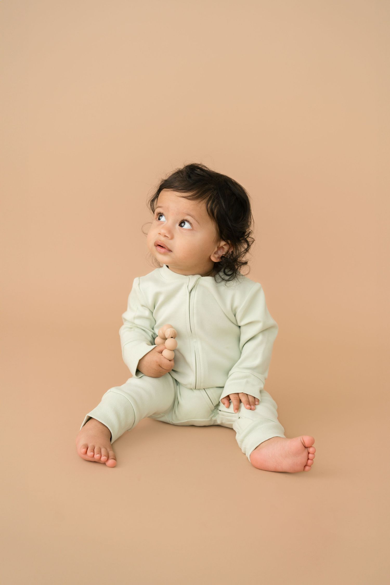 A small child in a mint green outfit sits on a neutral beige background looking upward.