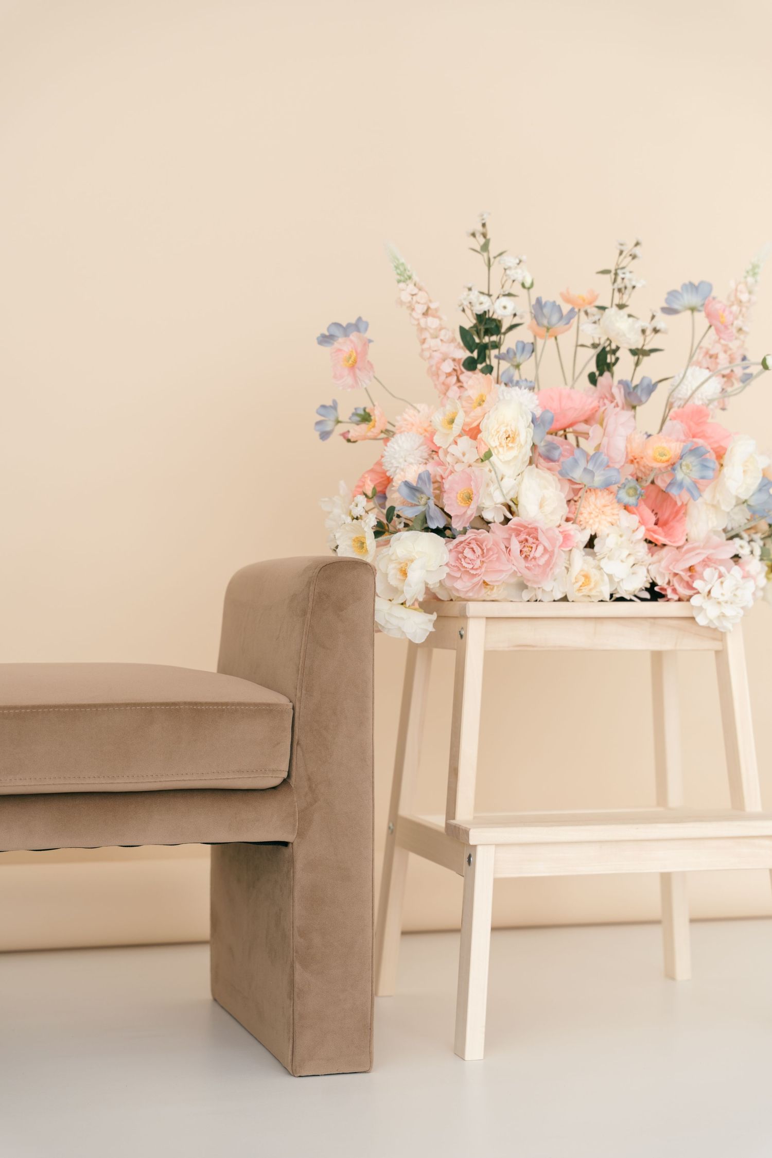 Elegant floral arrangement in pastel pink and blue sits atop white stepstool beside tan velvet sofa against beige wall.