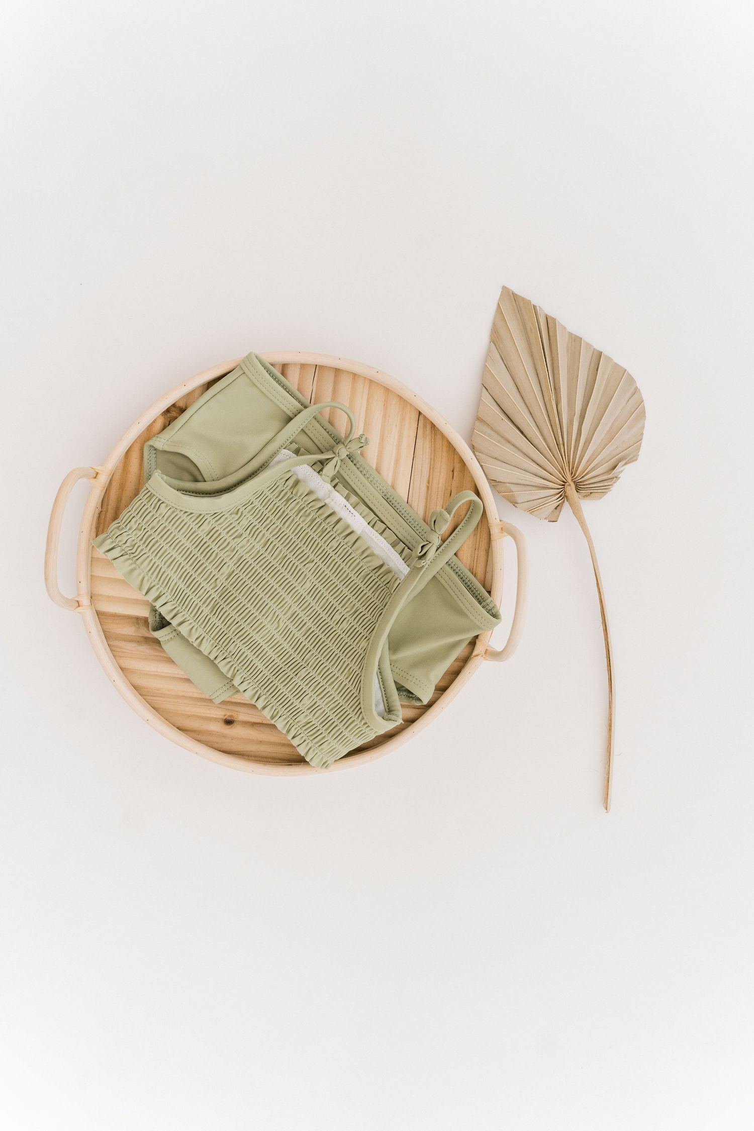 Sage green swimsuit on wooden tray with dried palm leaf decoration.