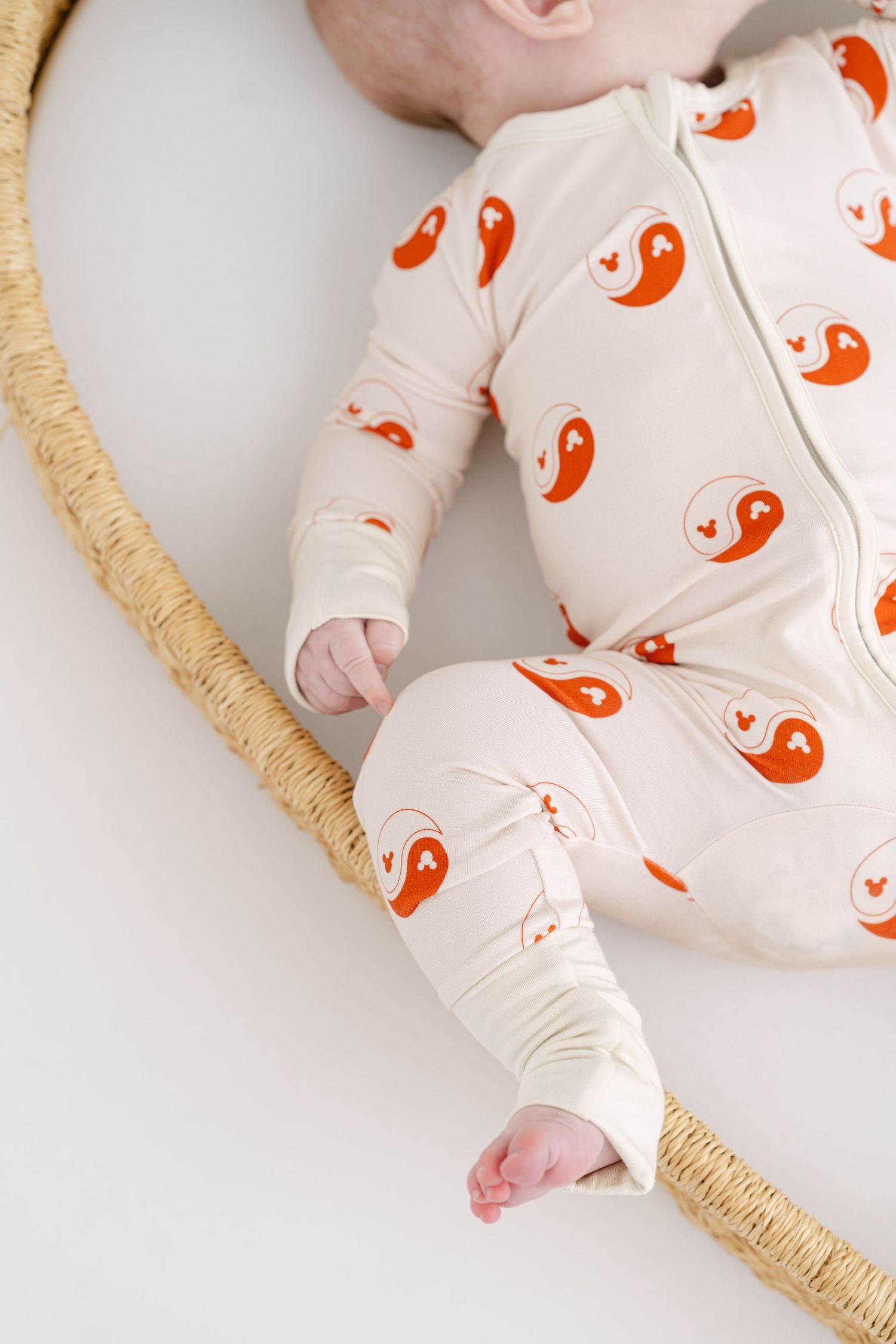 Baby wearing white sleeper with orange yin yang pattern lying on woven surface.