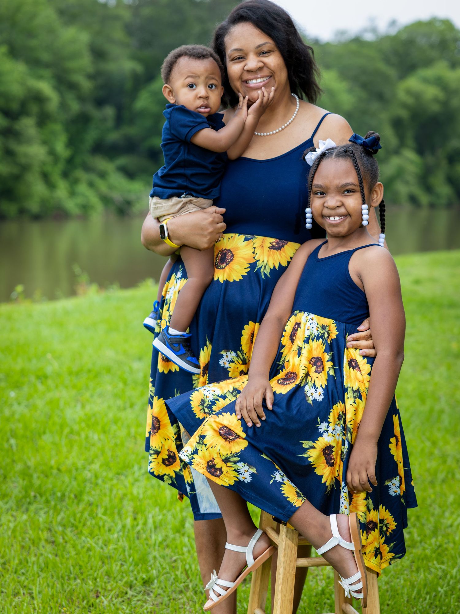 Navy blue and sunflower print coordinating dresses create a cheerful family portrait by a lake on green grass.