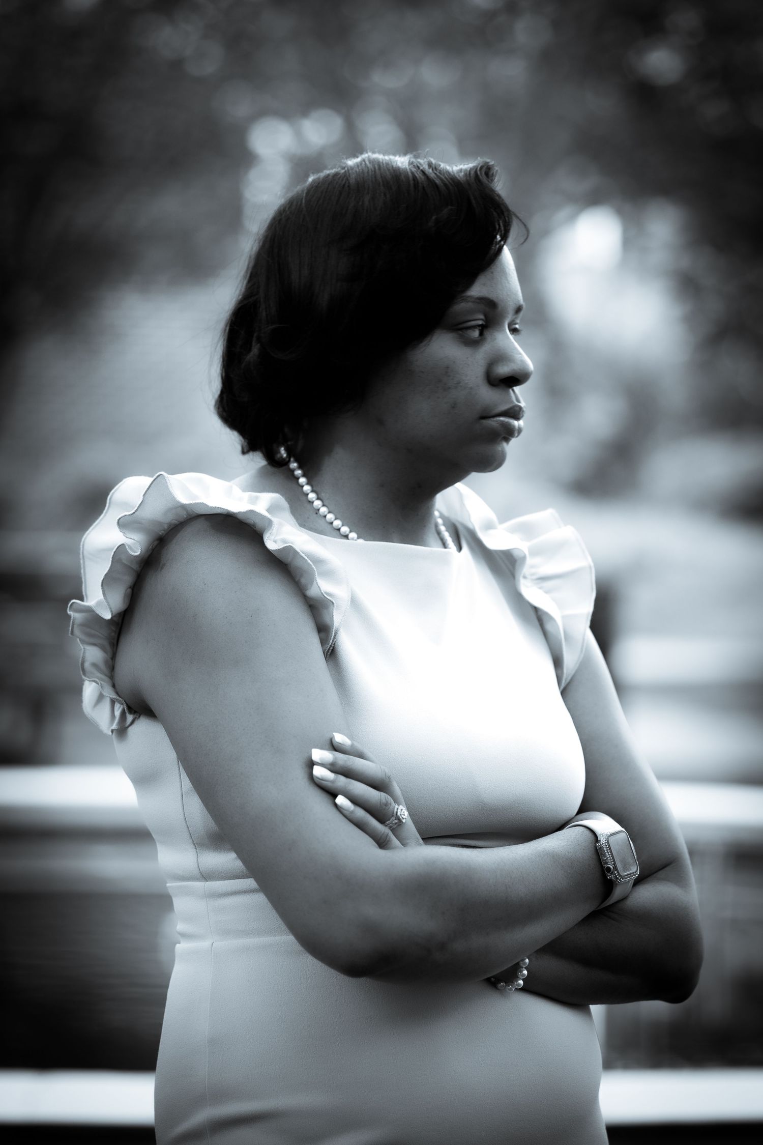 A black and white portrait of someone wearing a white dress with ruffles and pearl necklace, arms crossed in contemplation.