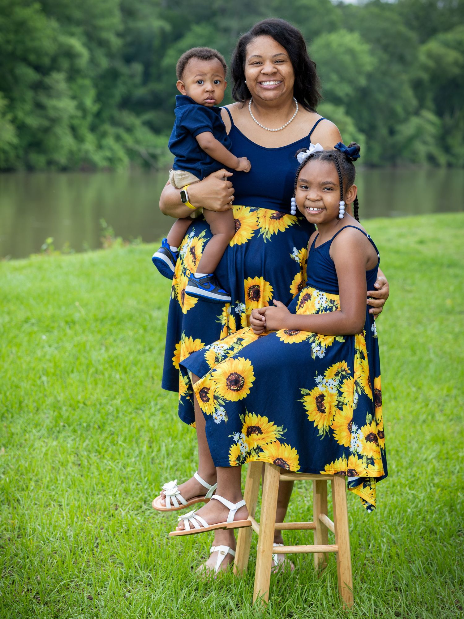 Beautiful family wearing coordinated navy blue and sunflower print dresses pose together by a lake on a sunny day.