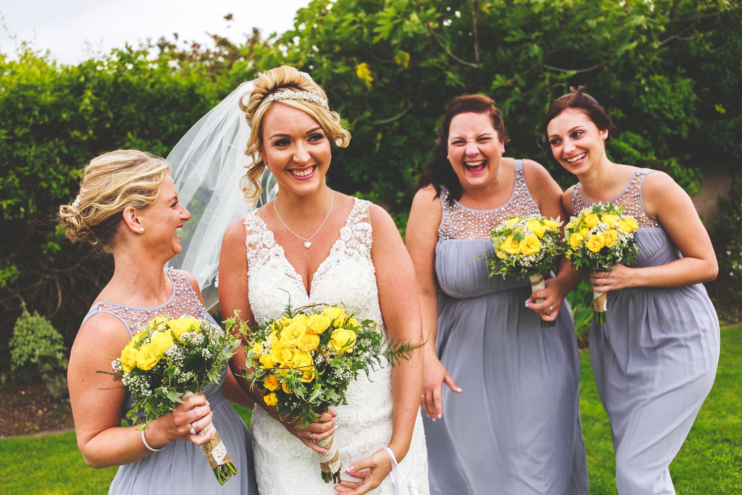 A bride in white dress and bridesmaids in grey dresses hold bright yellow bouquets while sharing a joyful moment.
