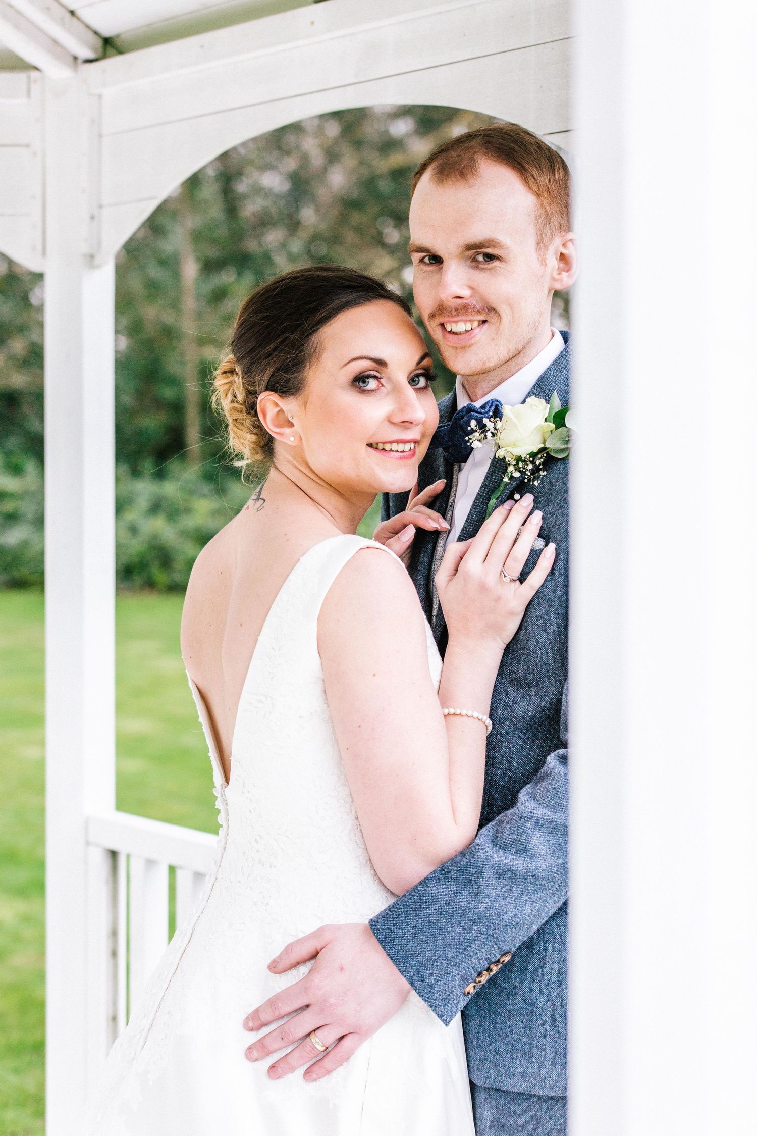 A romantic wedding photo of a couple posing in a white gazebo with greenery in the background.