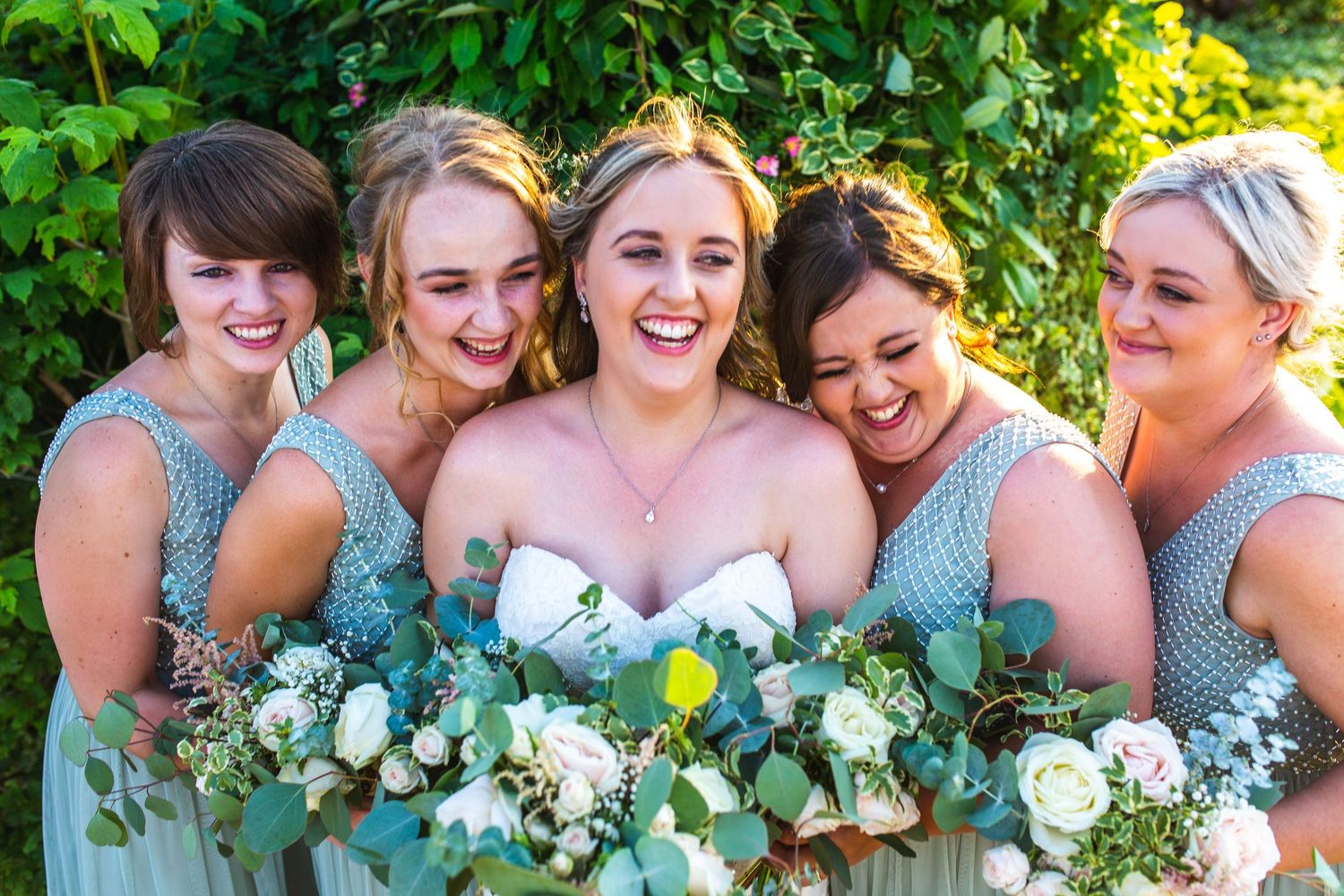 Wedding party in mint green bridesmaid dresses hold white and green floral bouquets while joyfully posing outdoors.