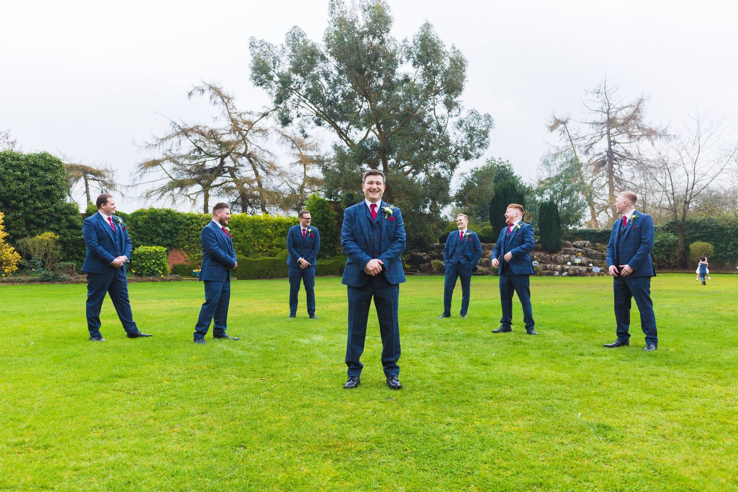 Wedding party in navy blue suits standing socially distanced on a green lawn with trees in the background.